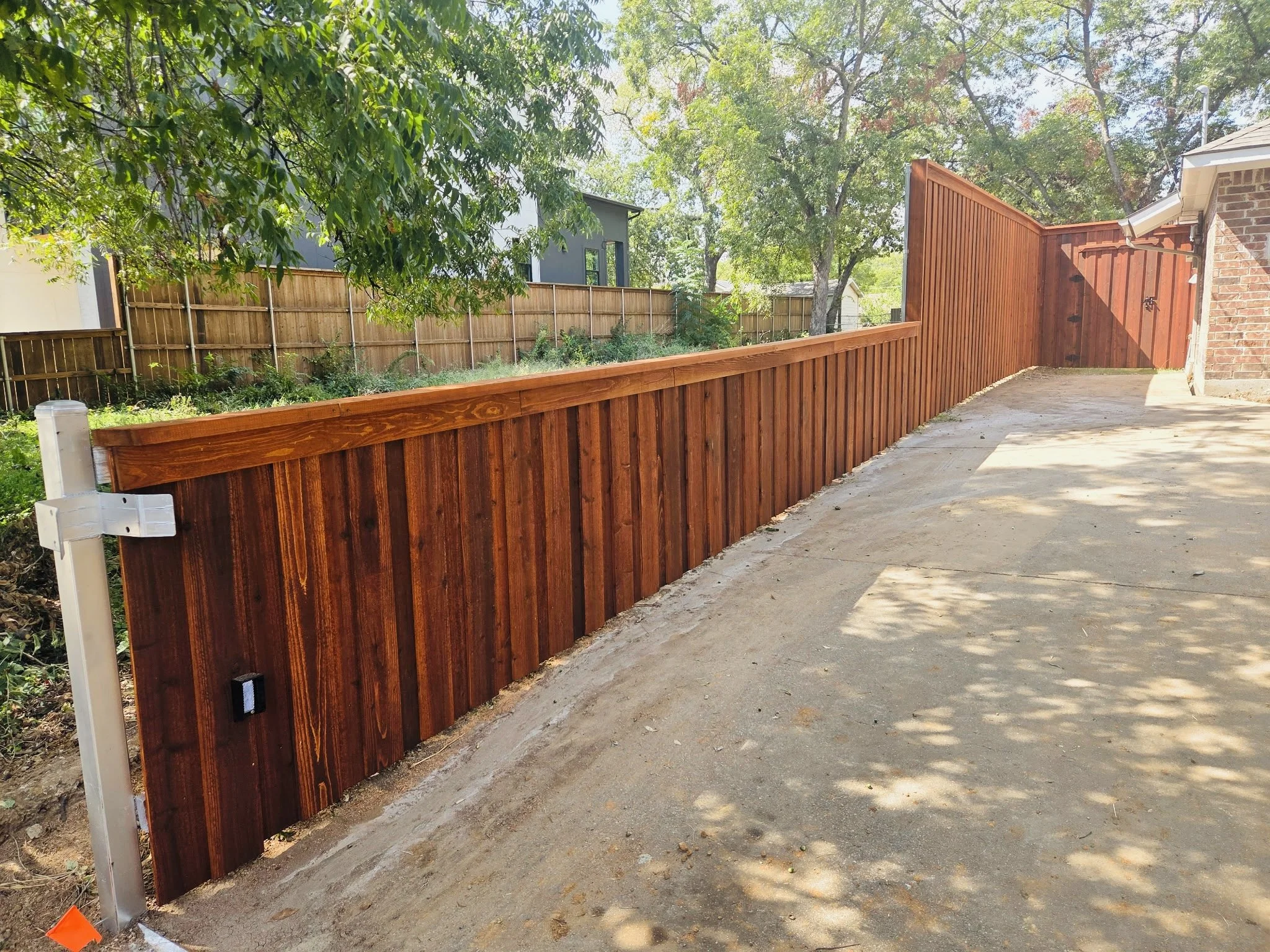 A backyard with a newly built wooden fence, trees with green leaves, and a concrete patio area. Shadows of trees are visible on the ground.