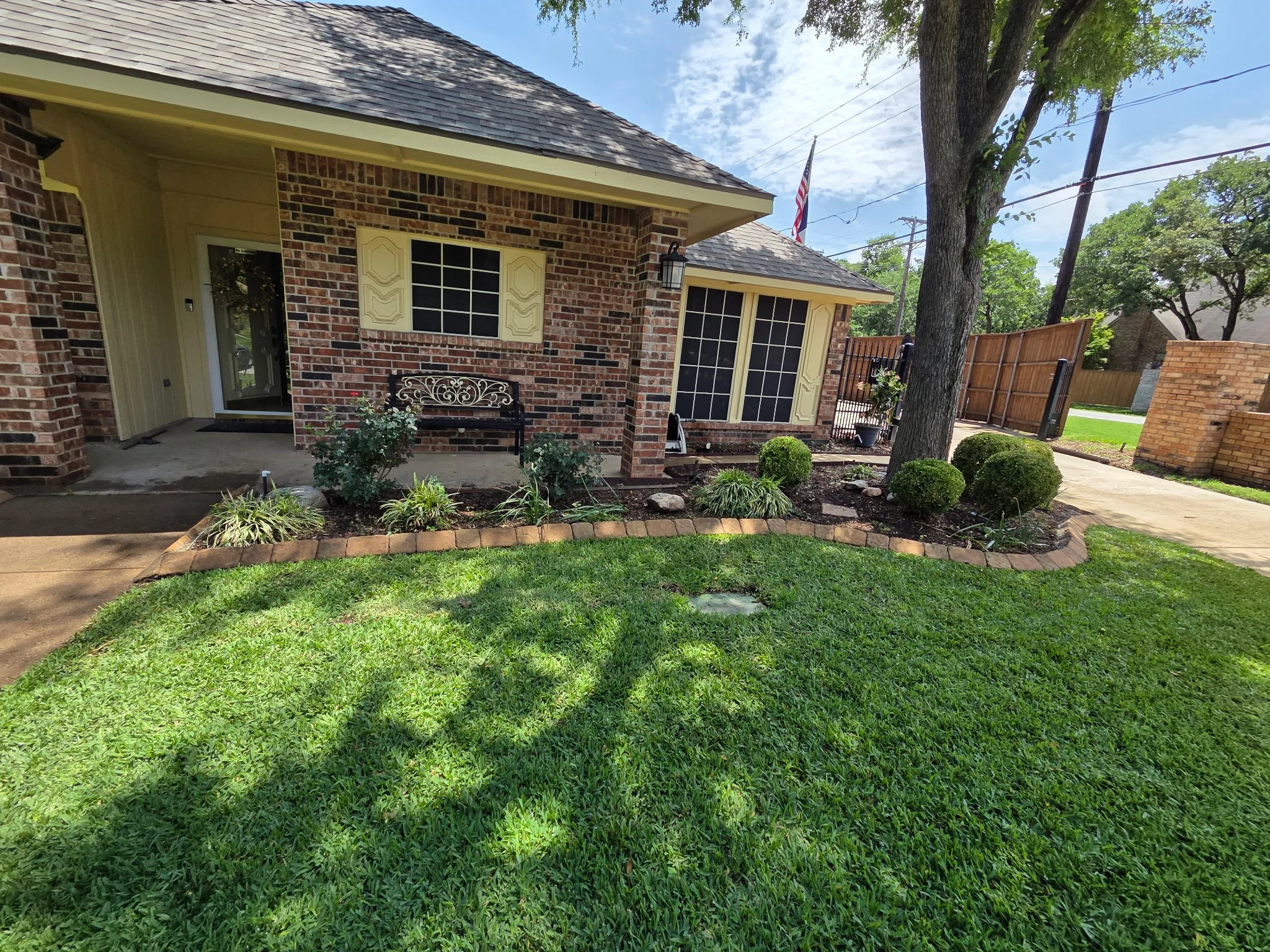 Front view of a brick house with a front porch, a black metal bench, and a landscaped garden with bushes and plants. Tall trees and a fence are visible in the background, along with an American flag.
