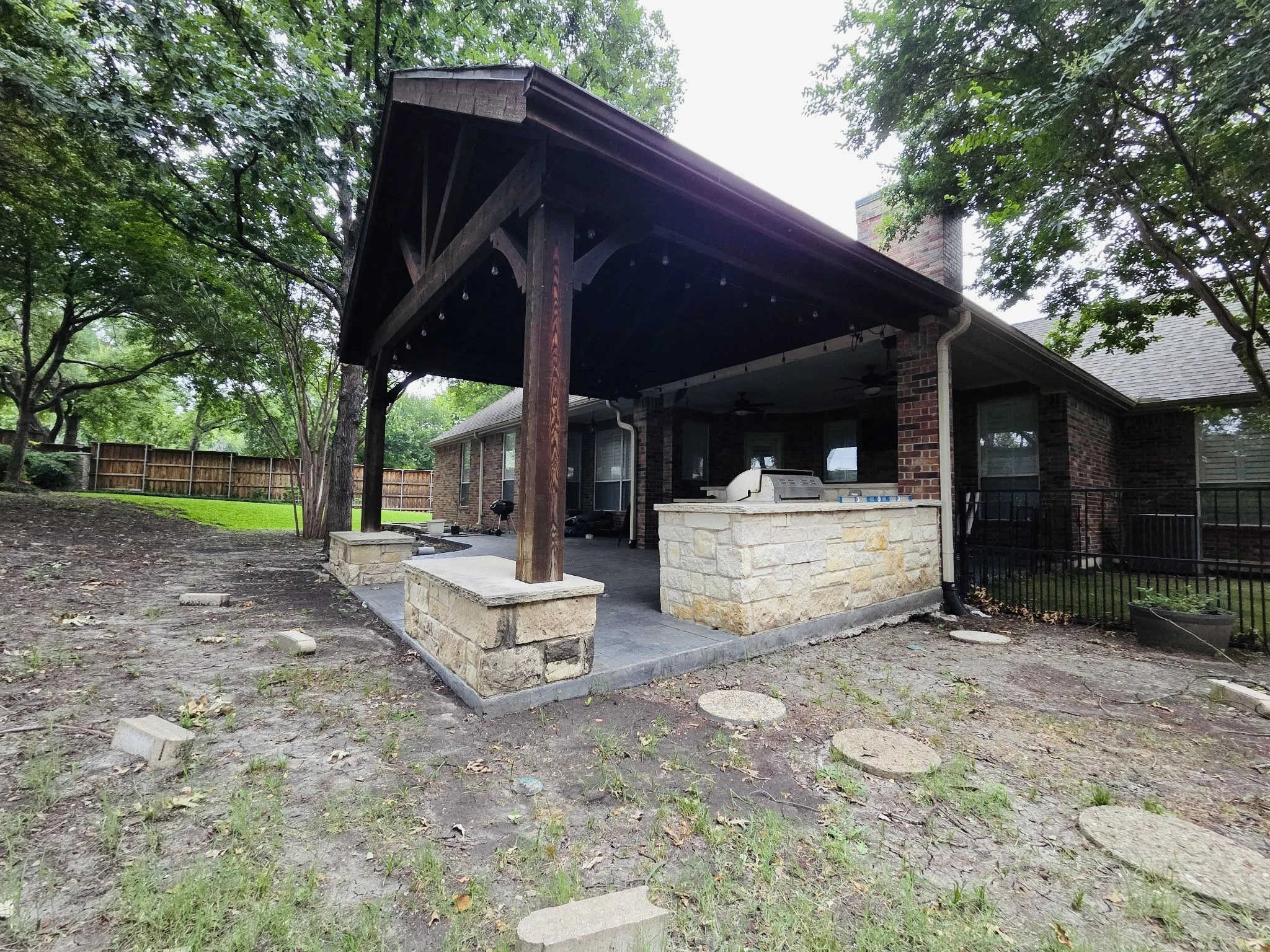 Backyard patio area with a covered porch, stone steps, grilling area, and a wooden fence in a green yard with trees.