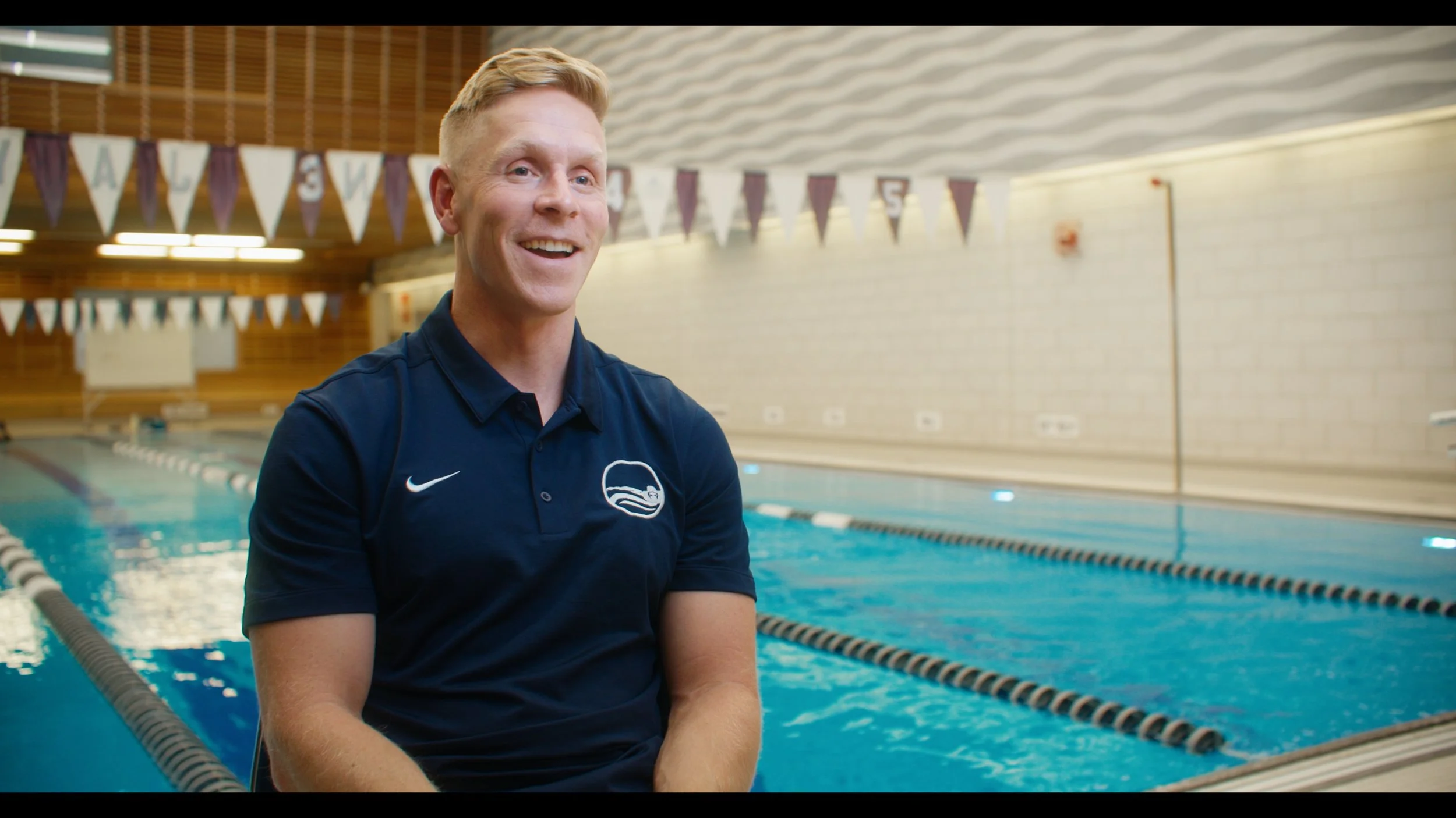 A smiling man in a navy blue polo shirt sitting poolside at an indoor swimming pool, with lanes marked by black and white lane dividers, and triangular flags hanging from the ceiling.