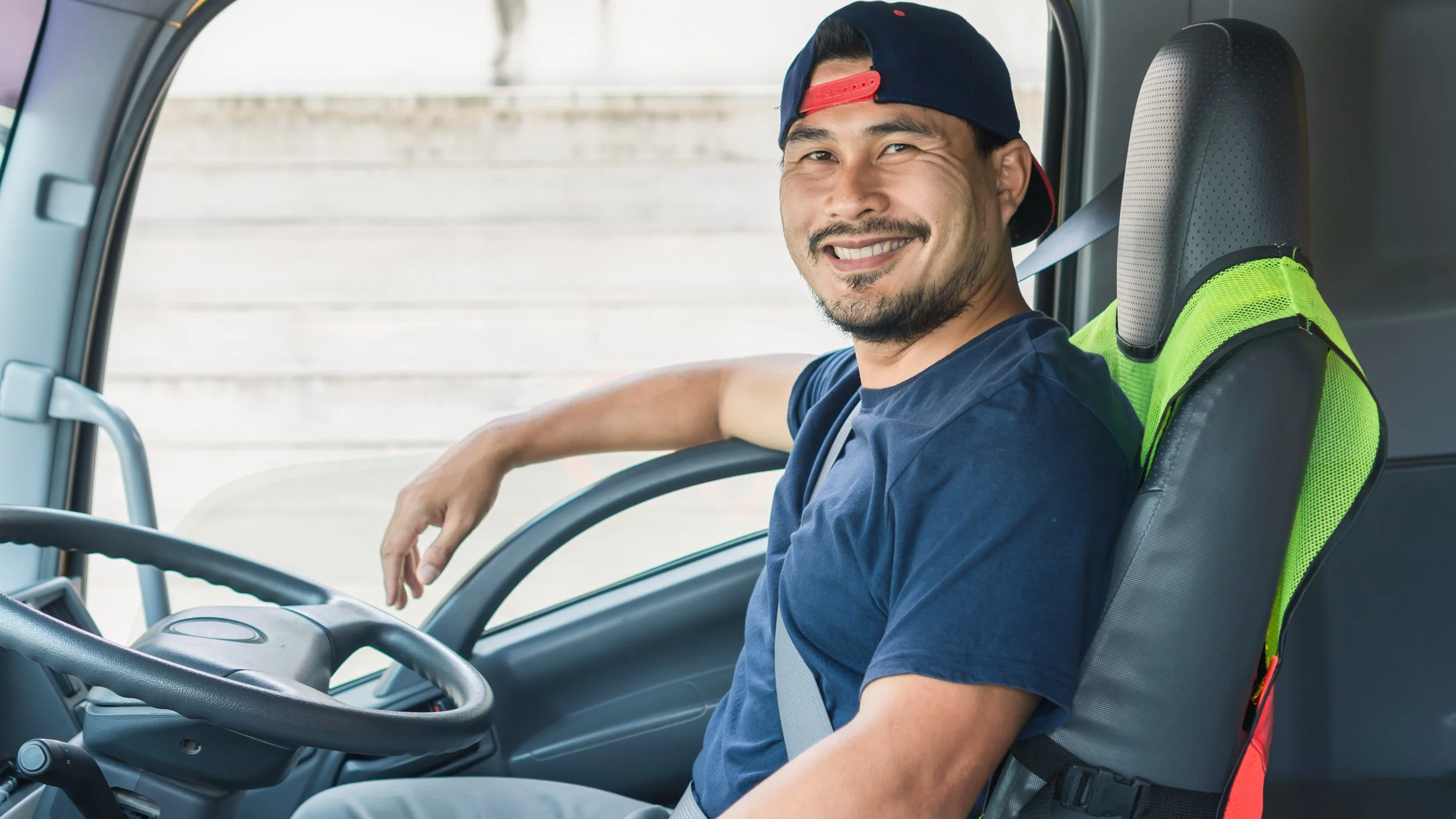A man sitting in the driver's seat of a truck or large vehicle, smiling, wearing a navy blue t-shirt and a backward baseball cap. A green safety vest is attached to the seat back.