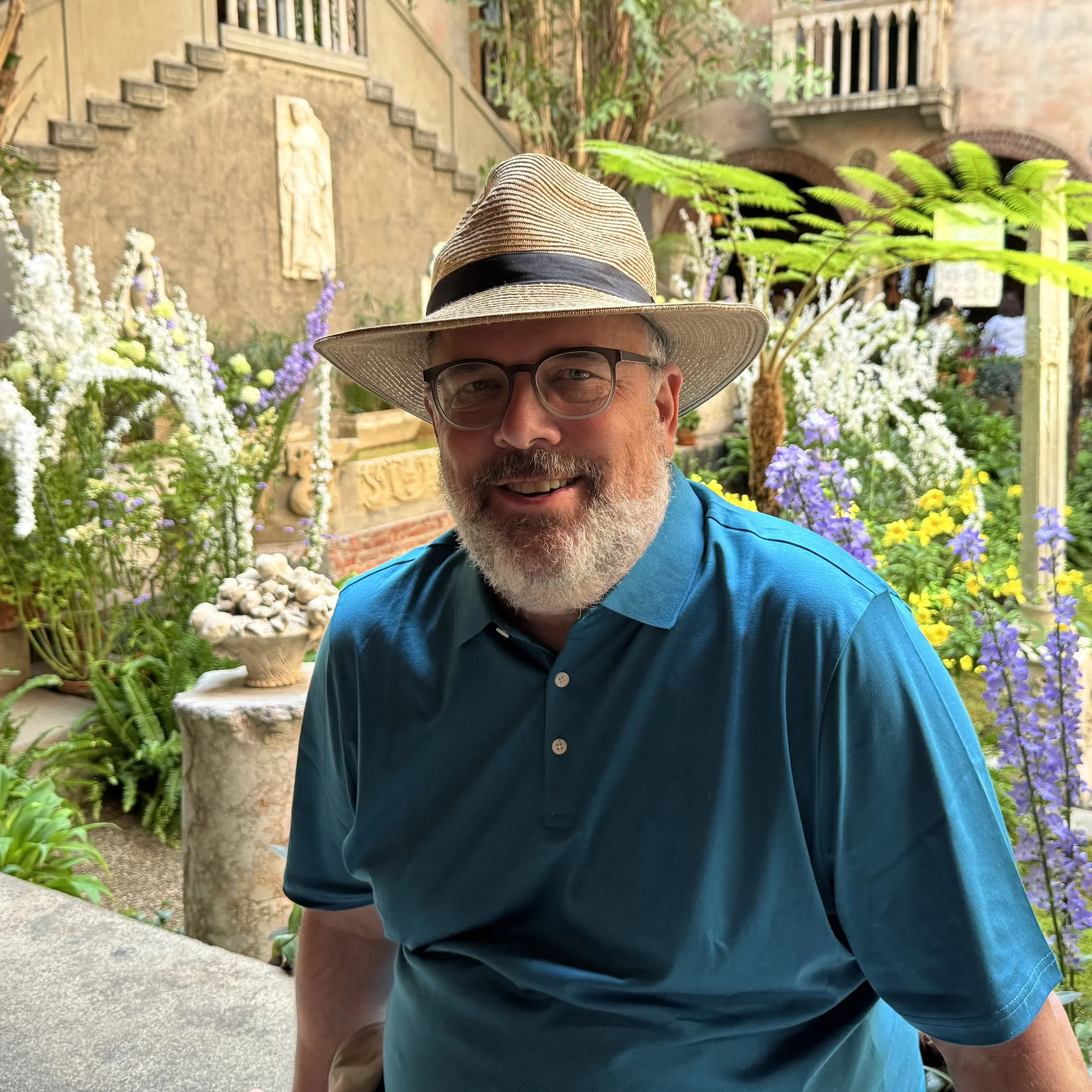 A smiling man with a beard, wearing glasses, a straw hat with a black band, and a blue collared shirt, standing in a lush garden with colorful flowers and greenery.