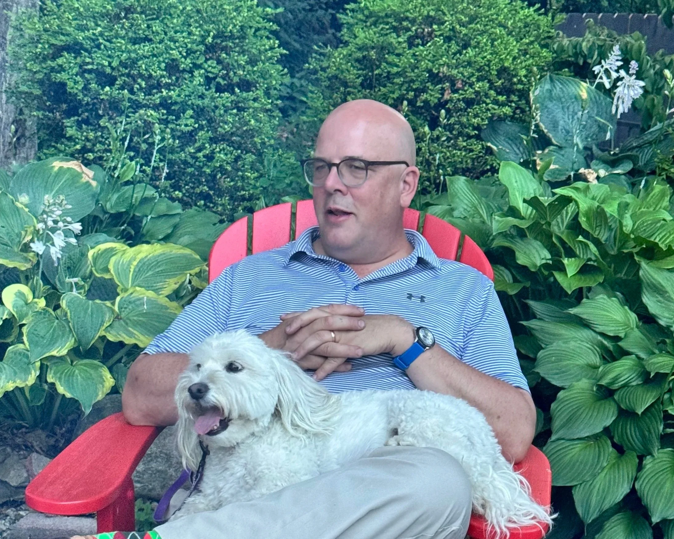 A man with glasses and a bald head sitting in a red chair outdoors, holding a white fluffy dog on his lap, surrounded by lush green plants.