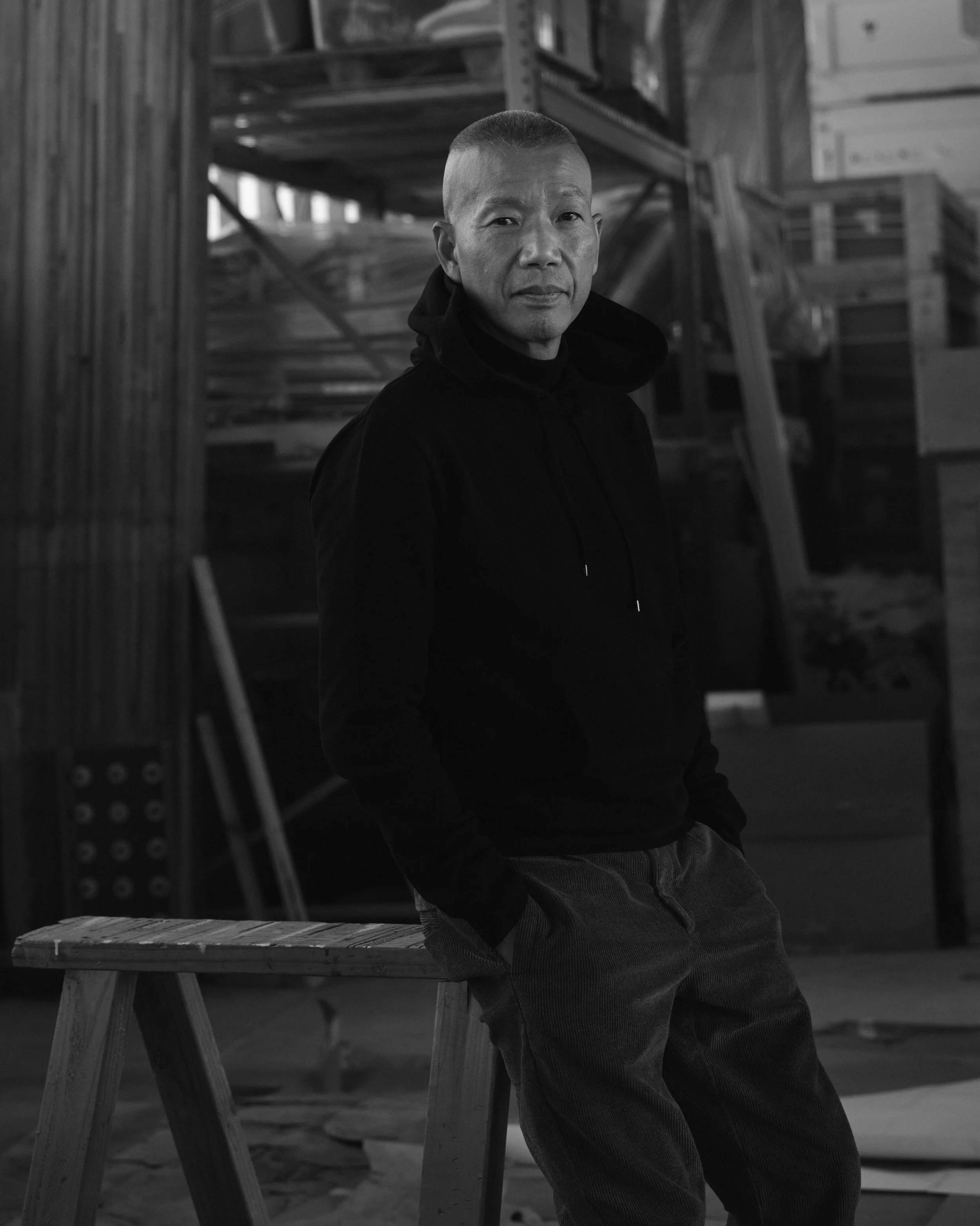 A man standing indoors with his hands in his pockets, wearing a black hoodie and pants, in front of wooden shelves and construction materials.