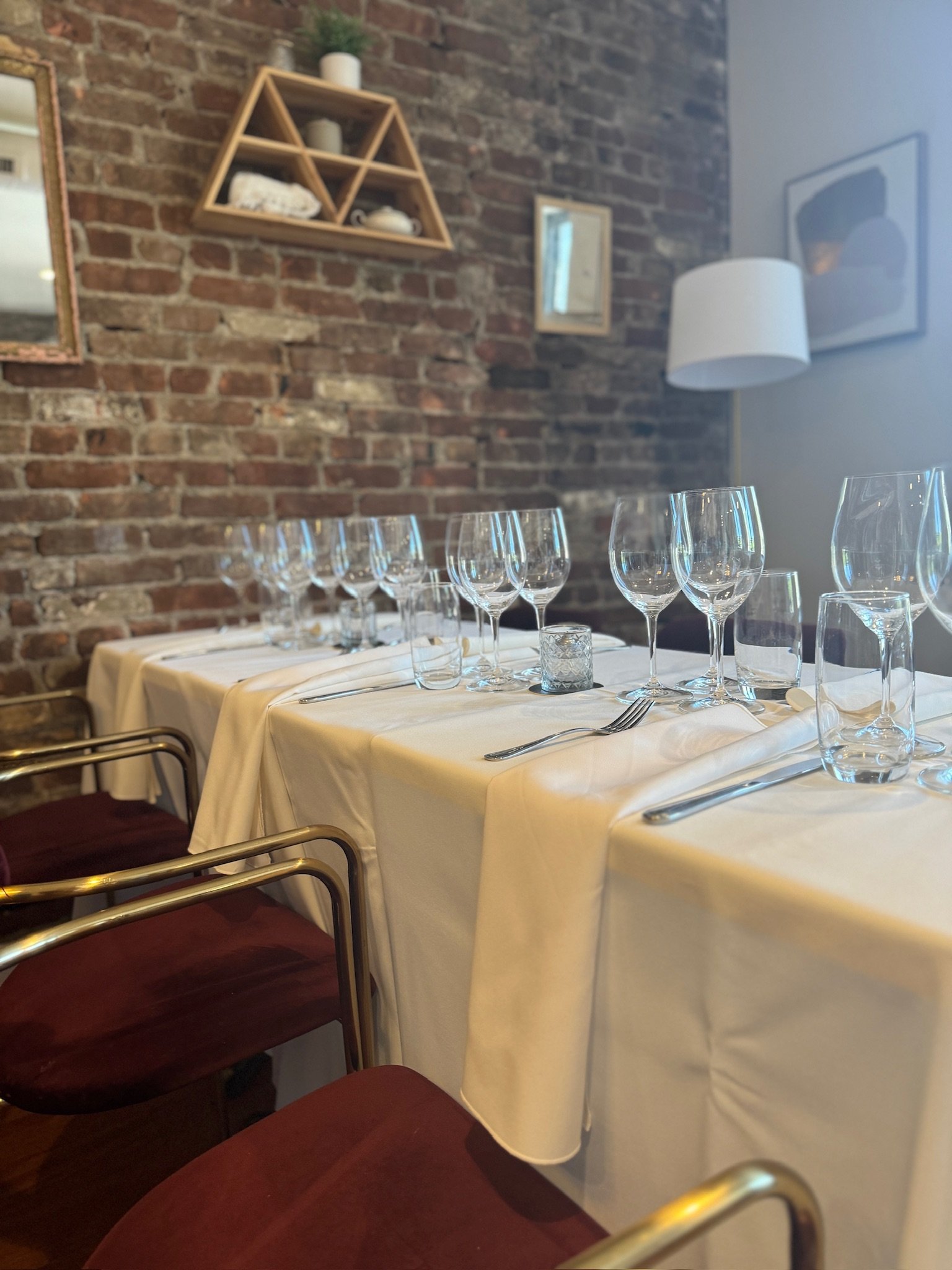 Elegant restaurant table set with white tablecloth, multiple empty wine glasses, water glasses, and silverware, against an exposed brick wall with decorative shelves, artwork, and a lamp.