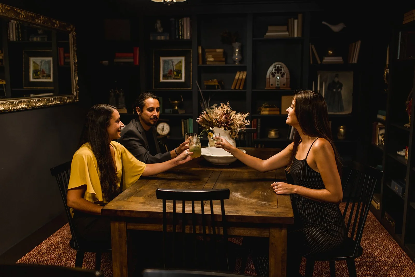 Four people sitting around a wooden table, two women and two men, are raising glasses in a toast in a dimly lit room with dark bookshelves and artwork in the background.