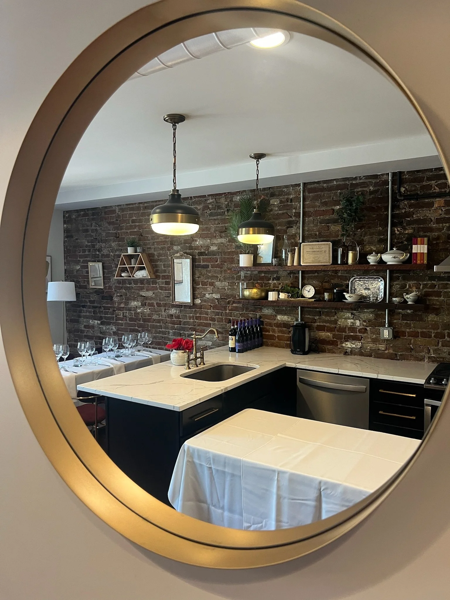 A modern kitchen viewed through a circular mirror. The kitchen features a brick wall with open shelves, pendant lights, a black island with a white marble countertop, and a small table set for dining.