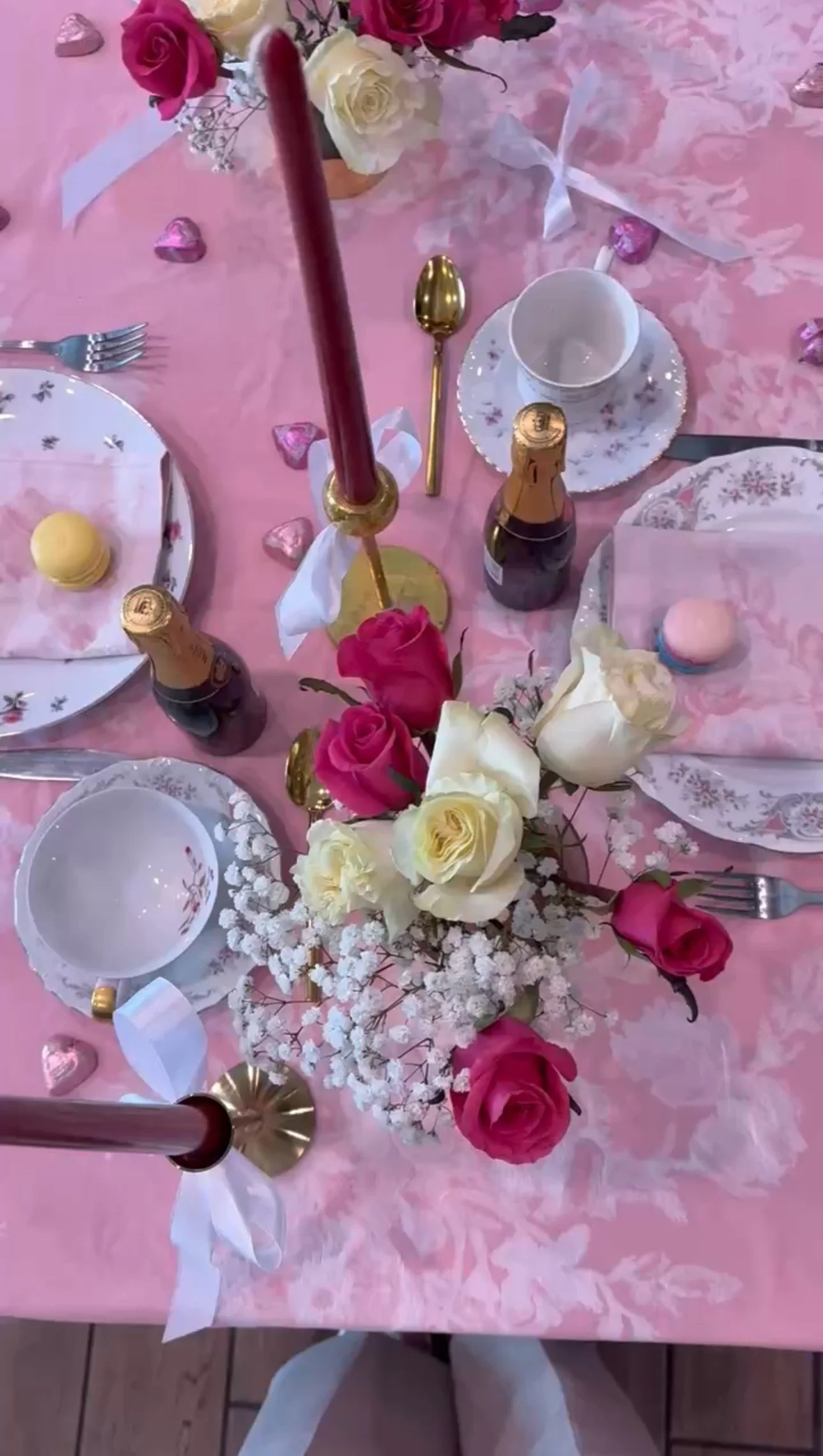 A table decorated for a celebration with pink and white roses and baby's breath floral centerpiece, pink tablecloth, pink heart-shaped chocolates, macarons, champagne bottles, gold and pink candles, and floral-patterned china and flatware.