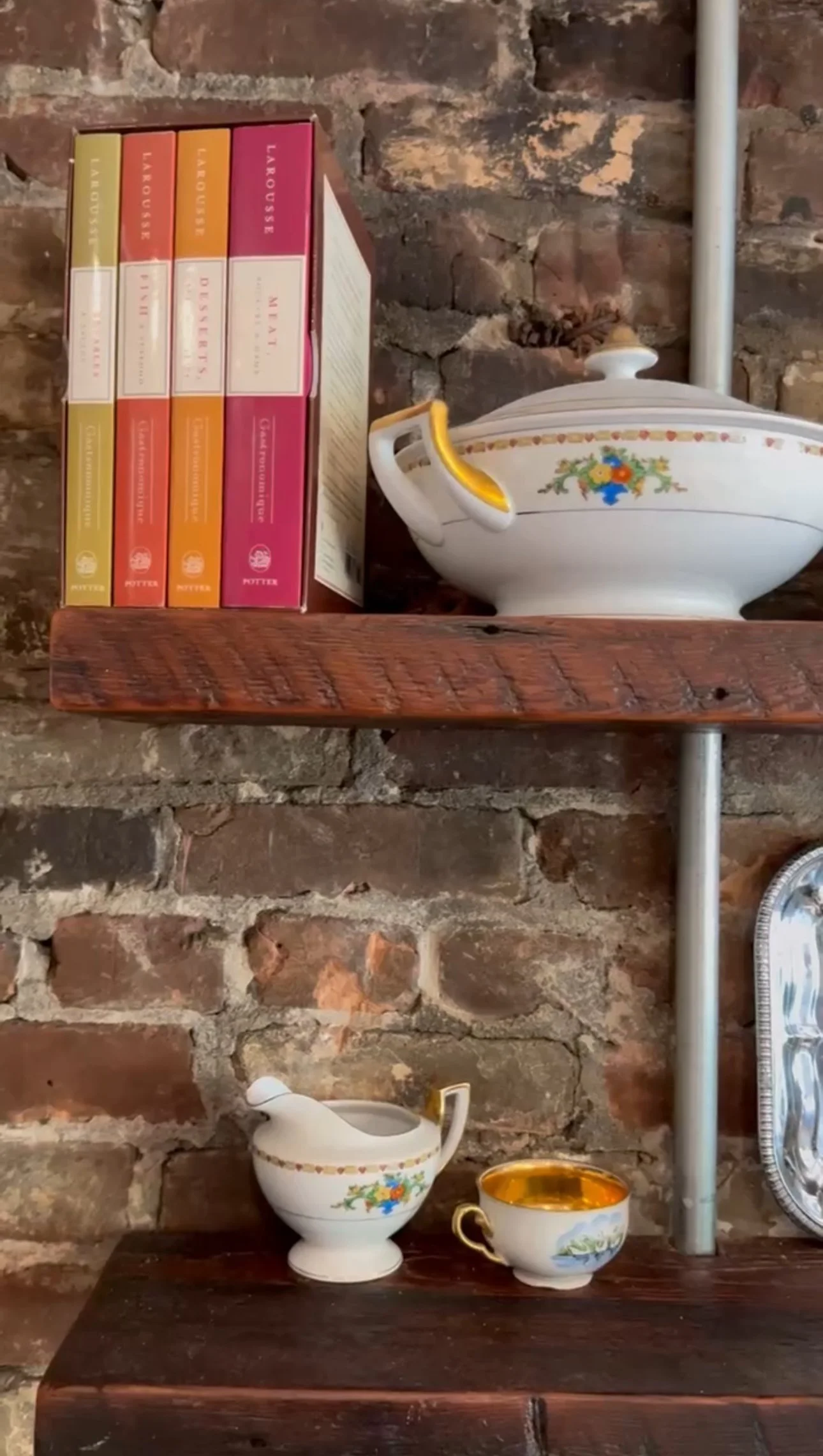 A rustic brick wall with wooden shelves holding vintage porcelain tea sets and books.