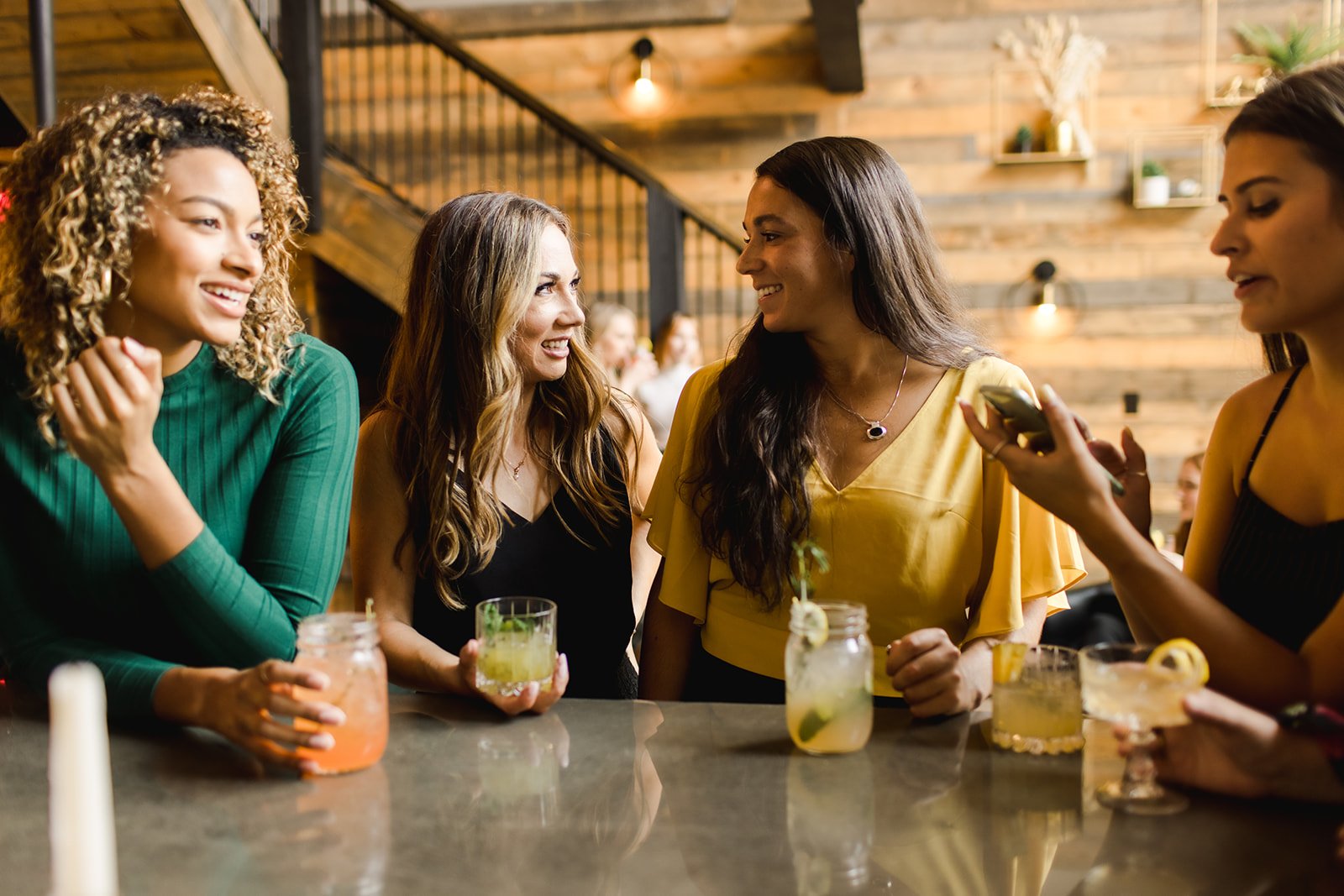 Four women are standing at a bar, engaging in conversation and smiling. They are holding glasses of colorful cocktails, and one woman is showing something on her phone. The background features warm, wood-paneled decor.