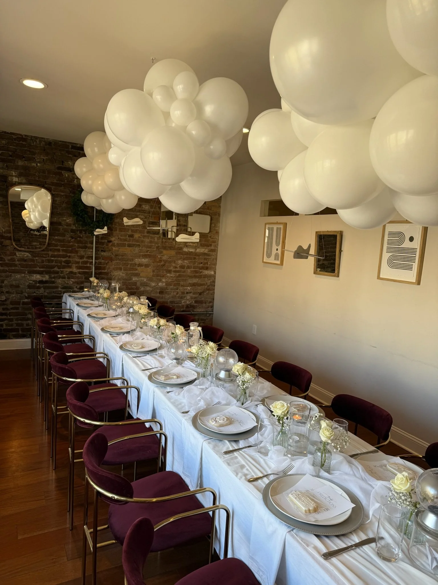 A decorated dining room with a long table set for a celebration, featuring white tablecloths, plates, utensils, and vases with white flowers. Large white balloons are arranged on the ceiling, and art frames adorn the walls.