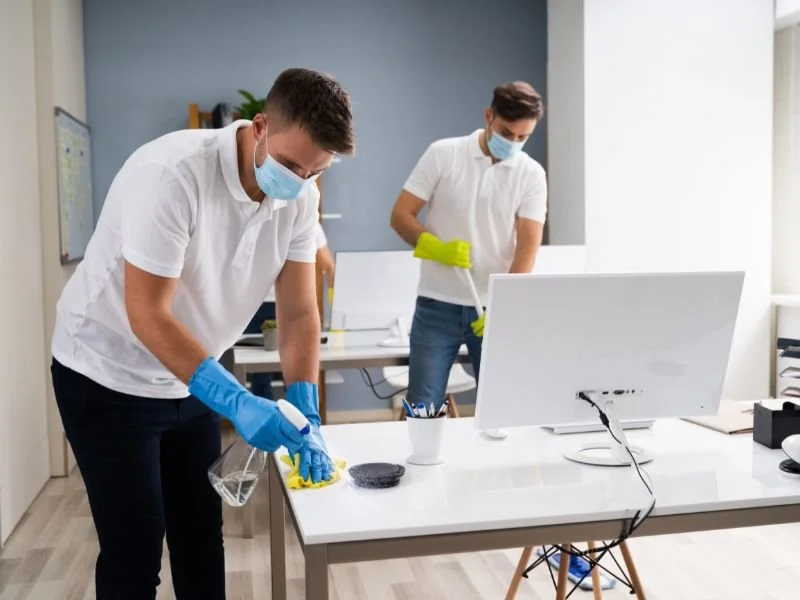 Two men wearing white shirts, protective masks, and gloves cleaning an office desk with cleaning supplies. One is wiping the desk with a cloth, while the other is using a mop.