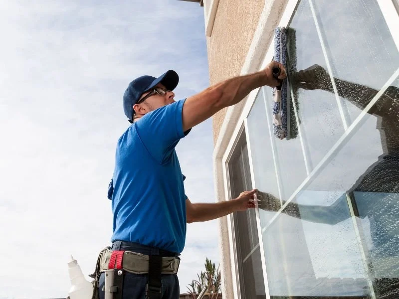 A man cleaning a large window with a squeegee outside a beige building on a partly cloudy day.