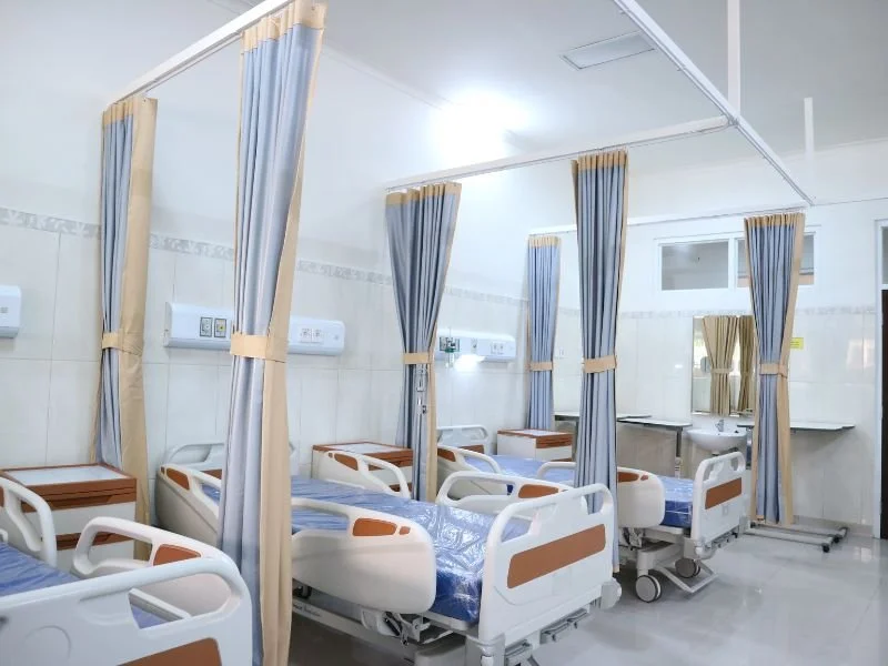 Hospital patient rooms with hospital beds separated by curtains, bedside tables, and medical equipment.