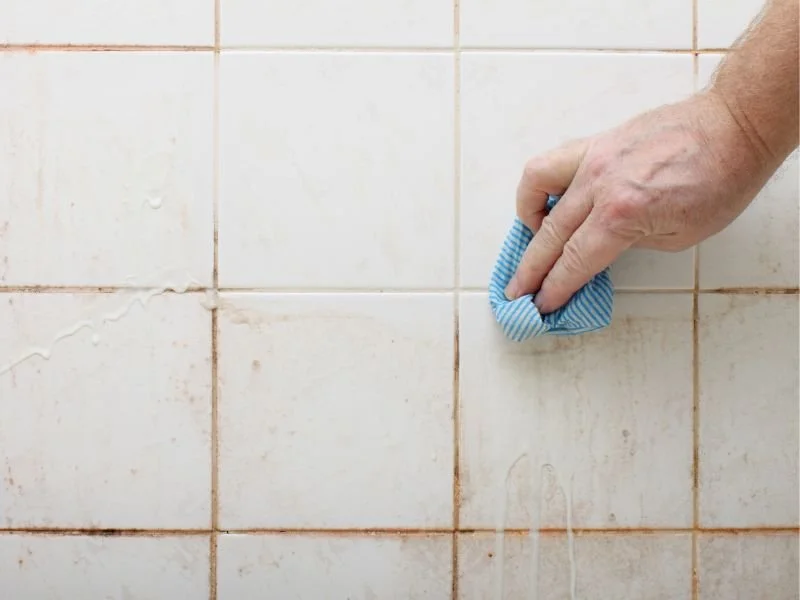 A hand cleaning dirty white tile wall with a blue and white striped cloth.