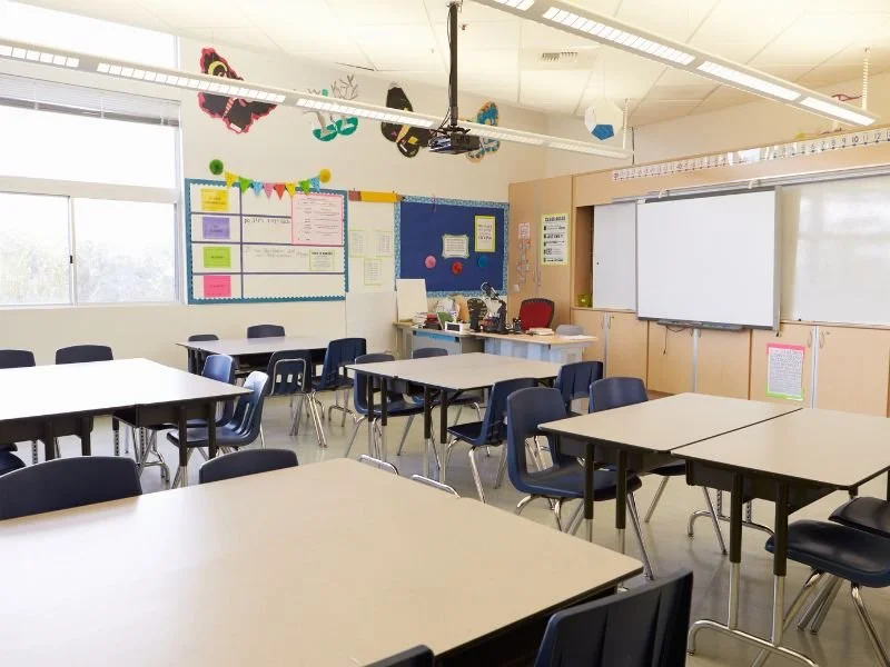 Empty classroom with white tables and navy chairs, large windows, and a whiteboard at the front. Decorations include a butterfly, colorful banner, and bulletin boards.
