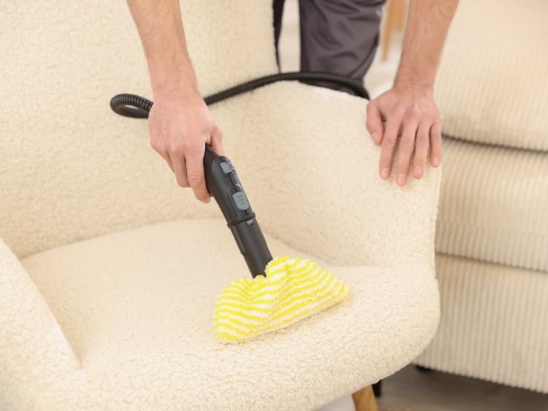 Person cleaning a beige sofa with a handheld vacuum and a yellow and white striped cloth.