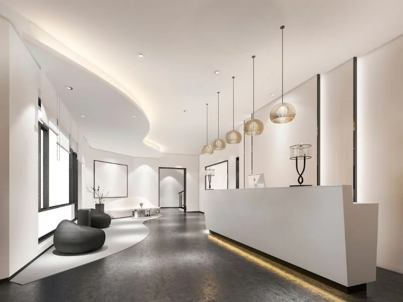 Modern, minimalist reception area with white walls, a curved ceiling, large windows, a white reception desk, pendant lights, a black sculpture on the desk, black seating, and decorative vases.