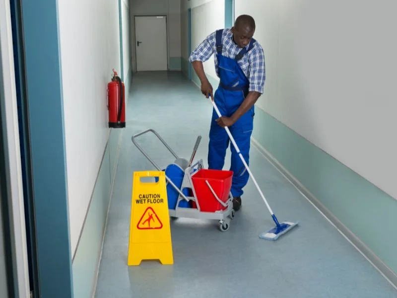 A man in blue overalls mops a hospital or facility corridor. A yellow wet floor caution sign is placed nearby, and cleaning supplies are on a rolling cart.