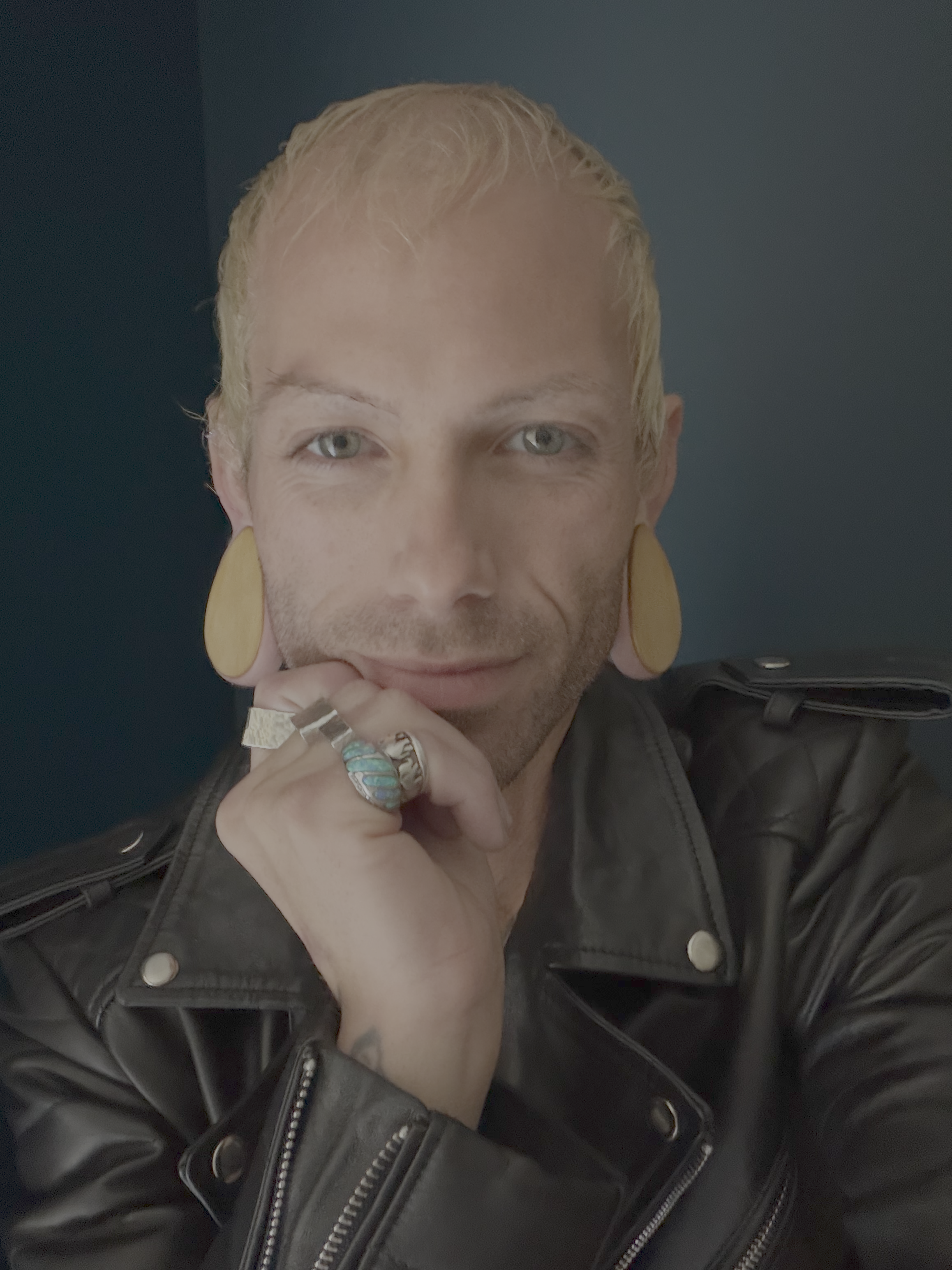 Close-up of a person with platinum blonde hair, wearing large yellow earrings, rings, and a black leather jacket, resting chin on hand against dark background.