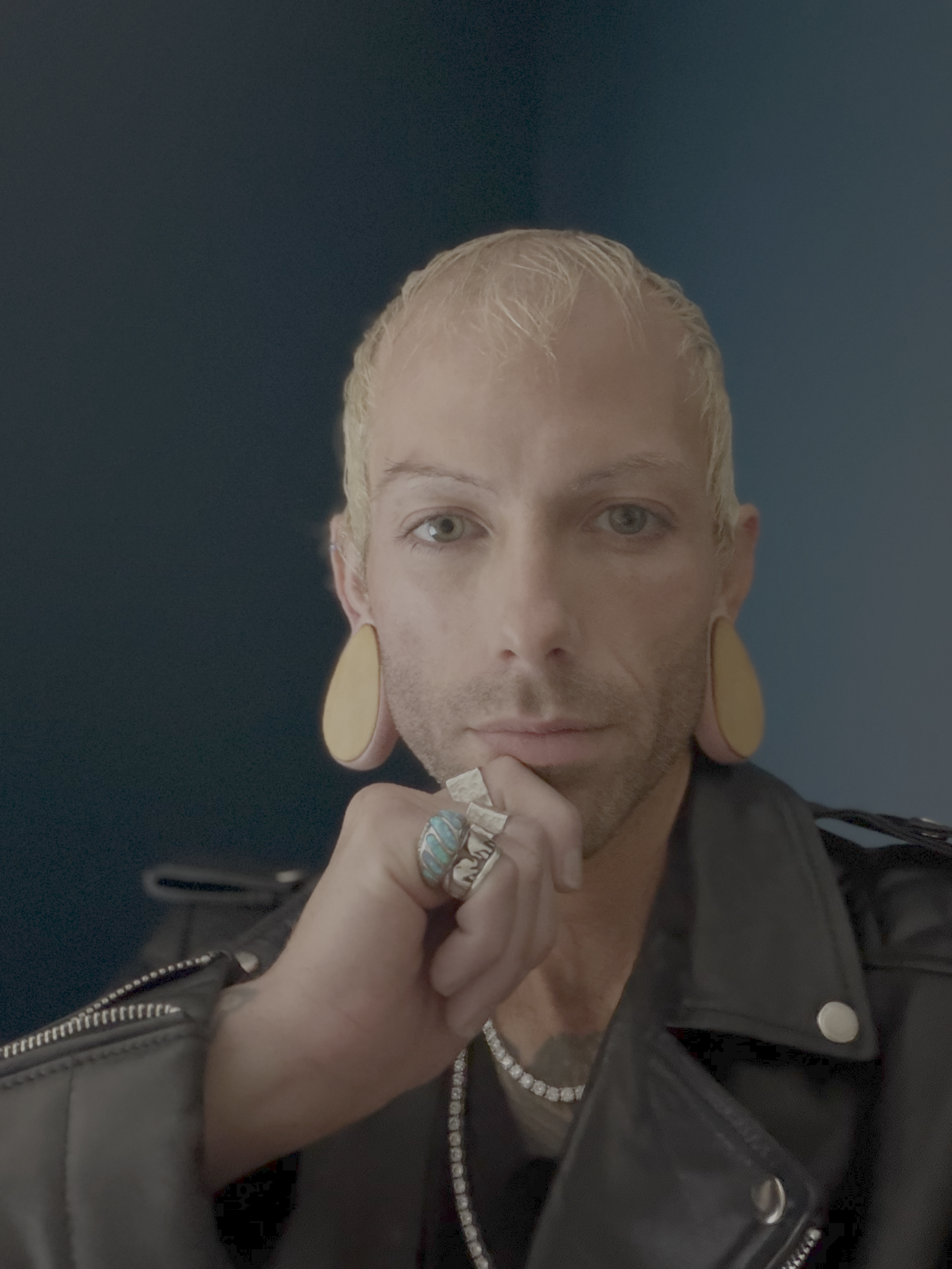 A person with short blonde hair and large earrings, resting their chin on their hand, wearing rings, necklaces, and a black leather jacket with a dark background.