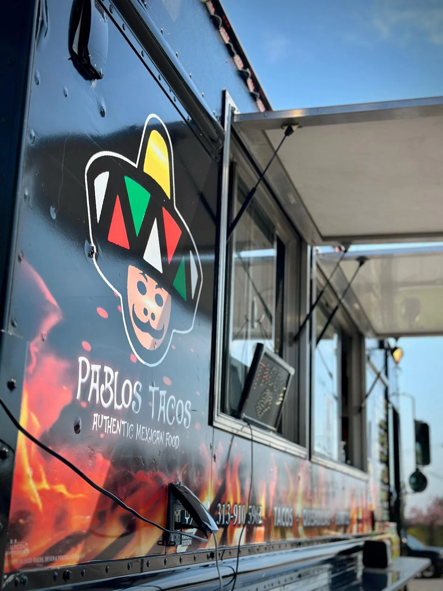 Side of a taco truck with a cartoon pig wearing a Mexican hat and colorful geometric patterns. Text on truck reads "PABLOS TACOS AUTHENTIC MEXICAN FOOD."