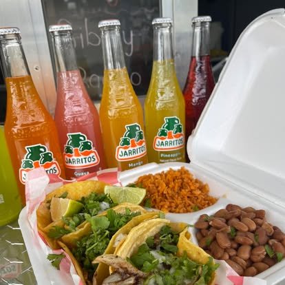 A tray with two tacos filled with shredded meat, lettuce, and lime wedges, served with side of pinto beans and Spanish rice, and four bottles of Jarritos soda in orange, pink, lemon-lime, pineapple, and strawberry flavors.