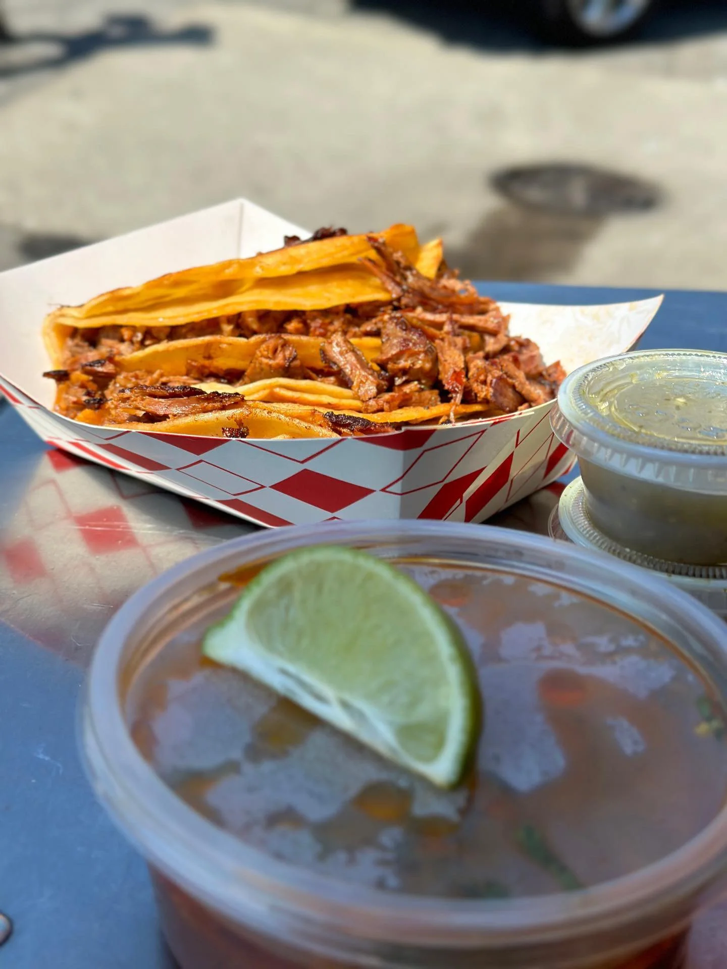 Close-up of a Mexican street food meal featuring tacos with shredded meat, a lime wedge, and small containers of salsa or sauces, on a metal table outdoors.