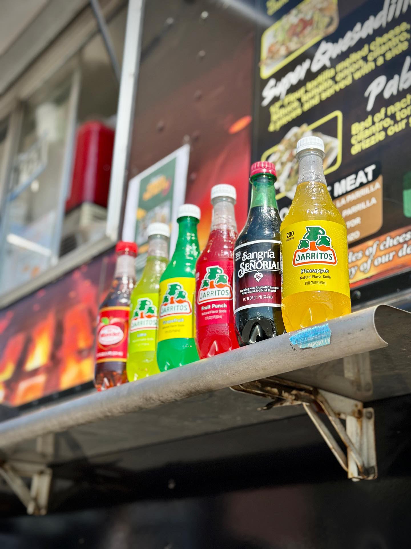 Seven bottles of soda on a metal shelf, including Jarritos pineapple, fruit punch, and mandarin flavors, as well as a Sangría Señorial non-alcoholic drink, with a black menu or sign in the background.
