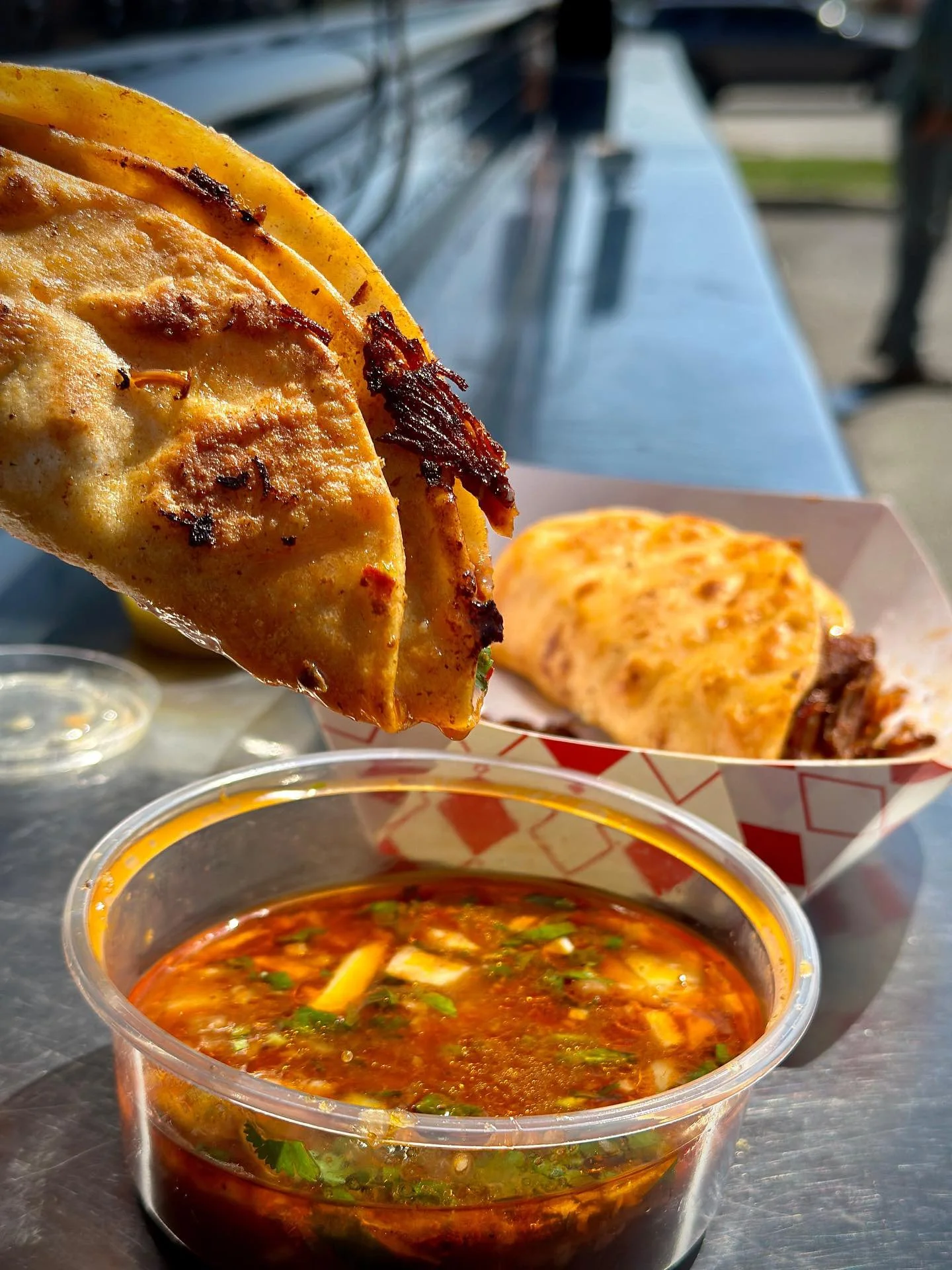 Close-up of a chicken taco with grilled chicken, on a tray with a plastic container of spicy salsa, and a burrito in a red and white checkered basket in the background.