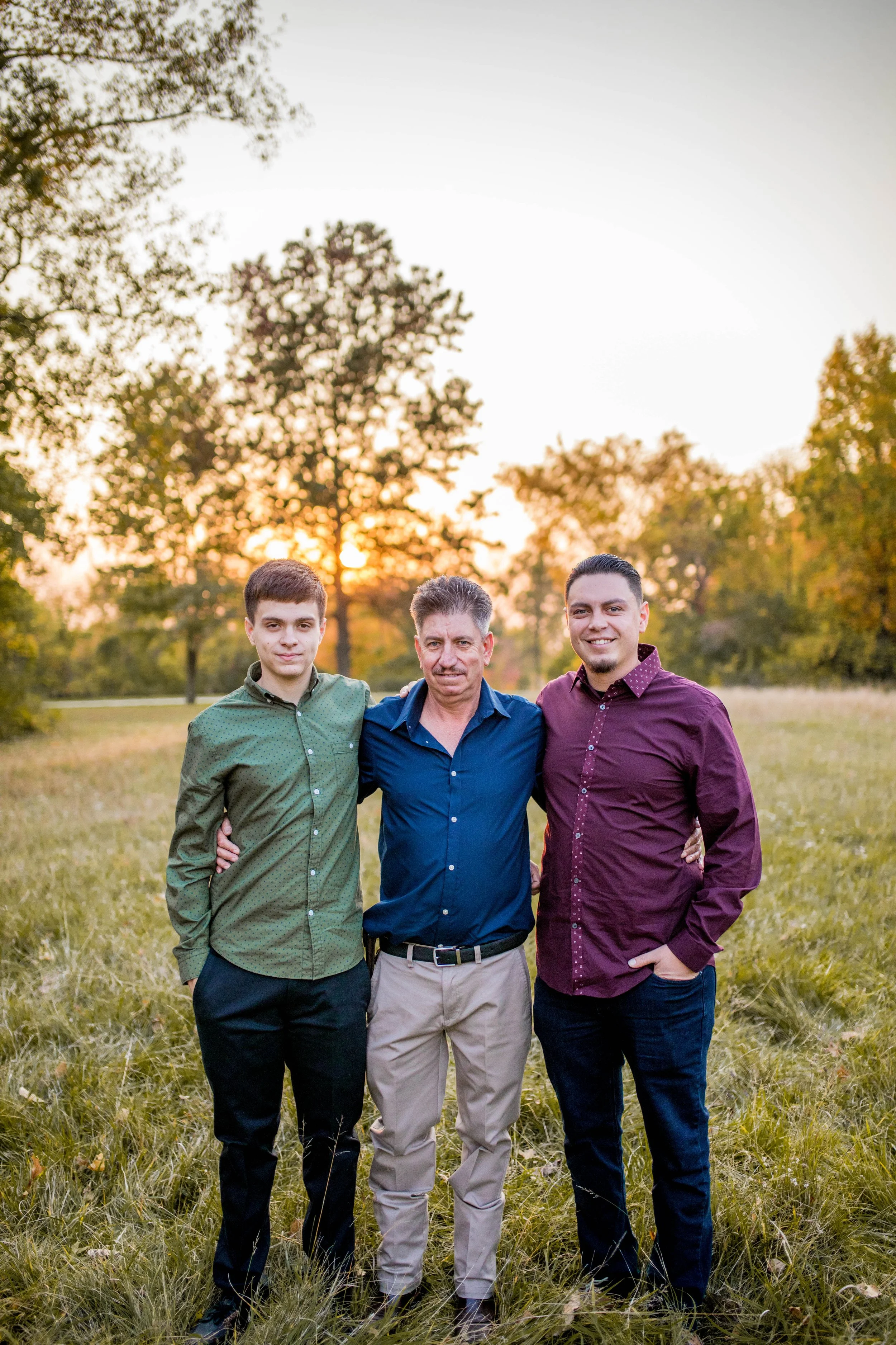 Three men standing together outdoors during sunset in a grassy field with trees in the background.