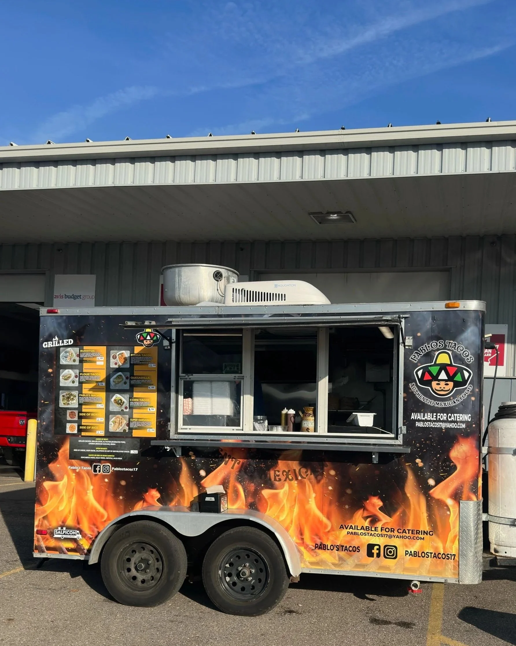 Food truck with flaming design, menu on the side, serving window open, parked in lot under blue sky.