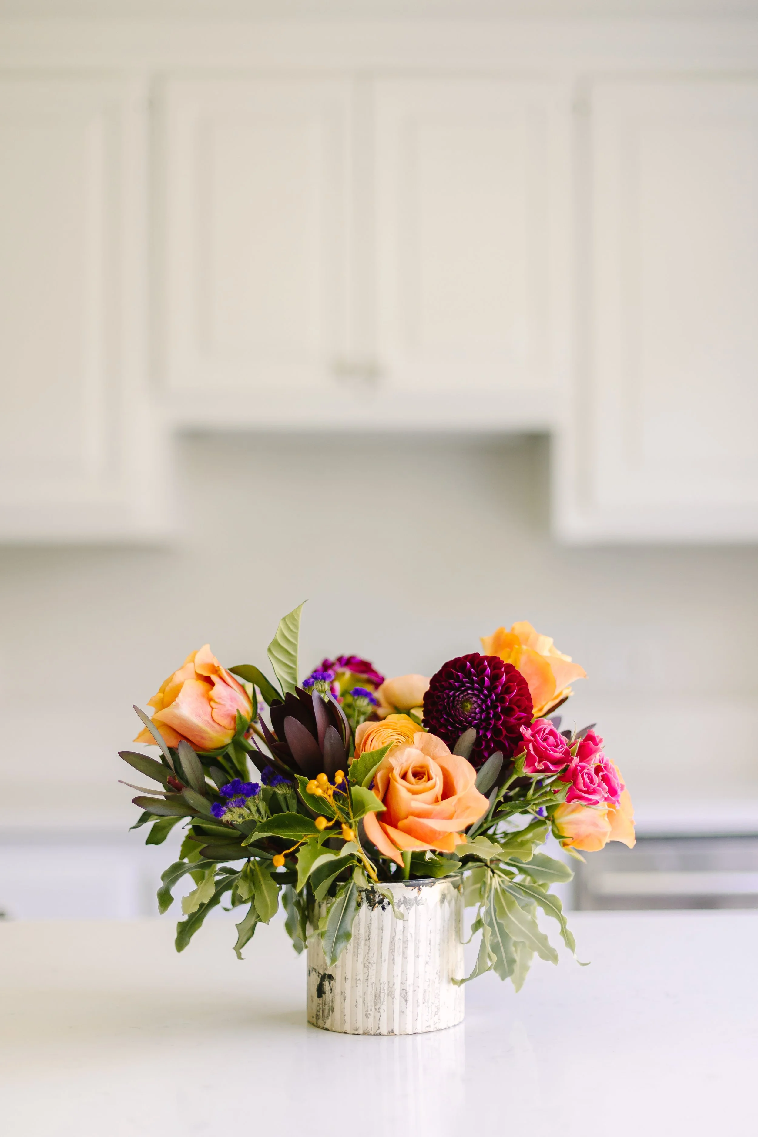 A vibrant flower bouquet with roses, dahlias, and greenery in a white ceramic vase on a light surface, with a white kitchen cabinets background.