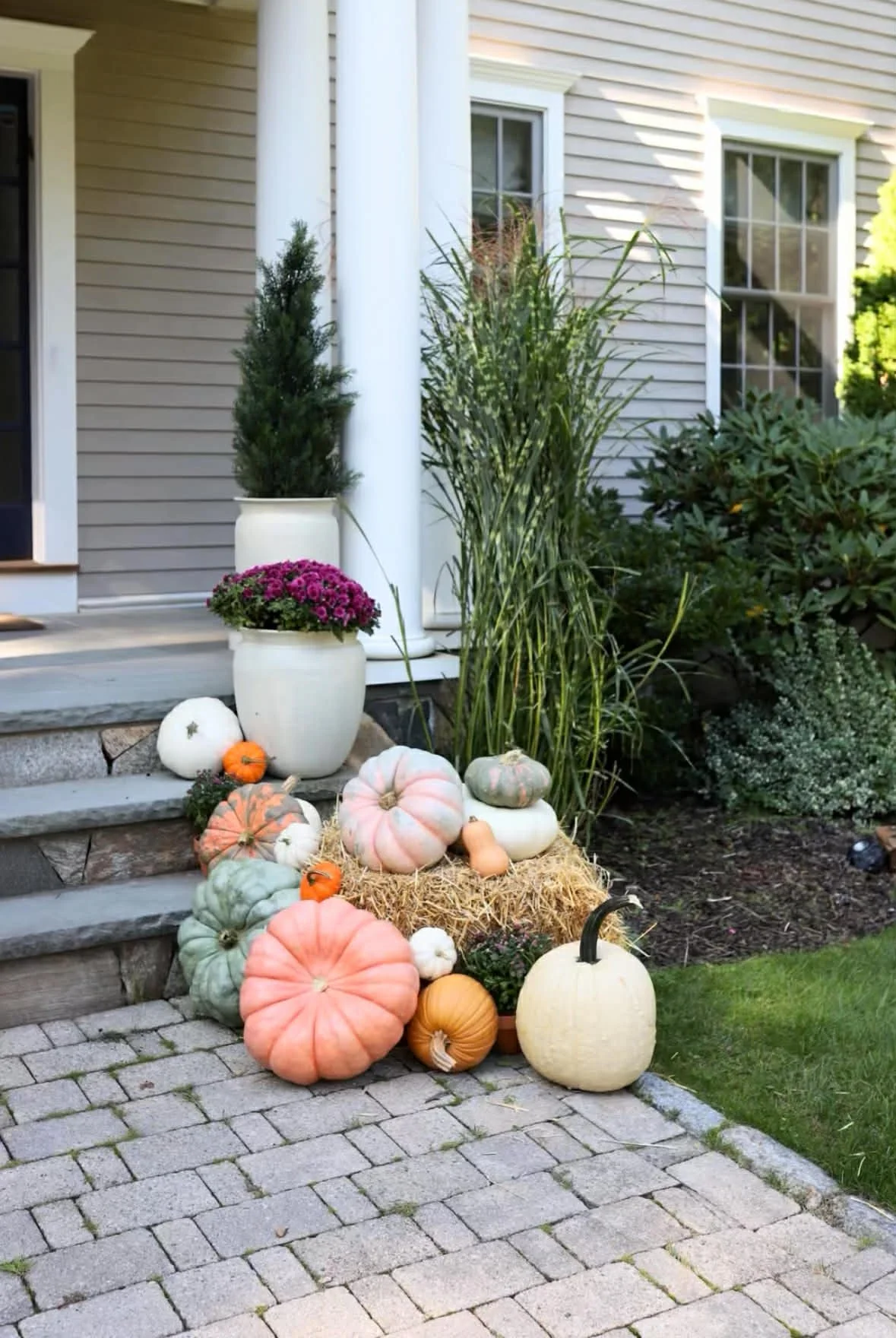 Fall harvest display with pumpkins, gourds, potted plants, and hay bales near a Wellesley home with beige siding and white columns.