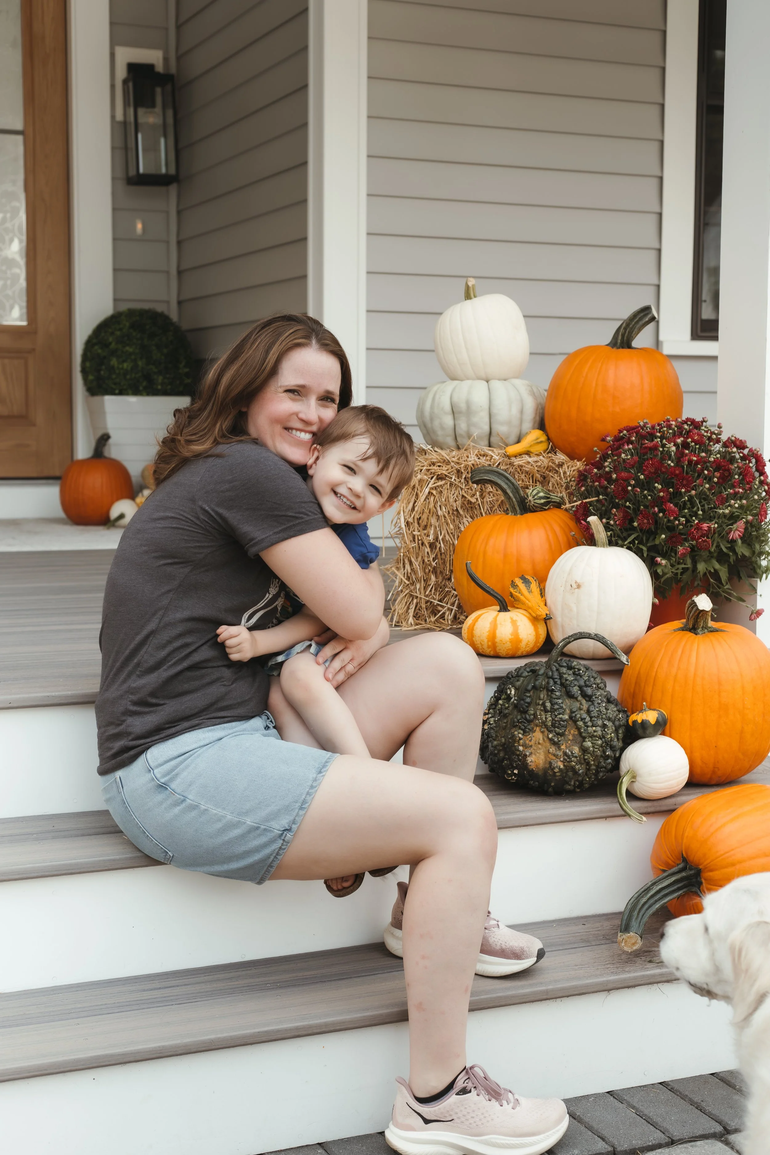 A woman and a young boy sitting on the front steps of a house, smiling and hugging each other. Around them, there are various pumpkins and gourds, fall flowers, and hay bales, creating a festive autumn display.