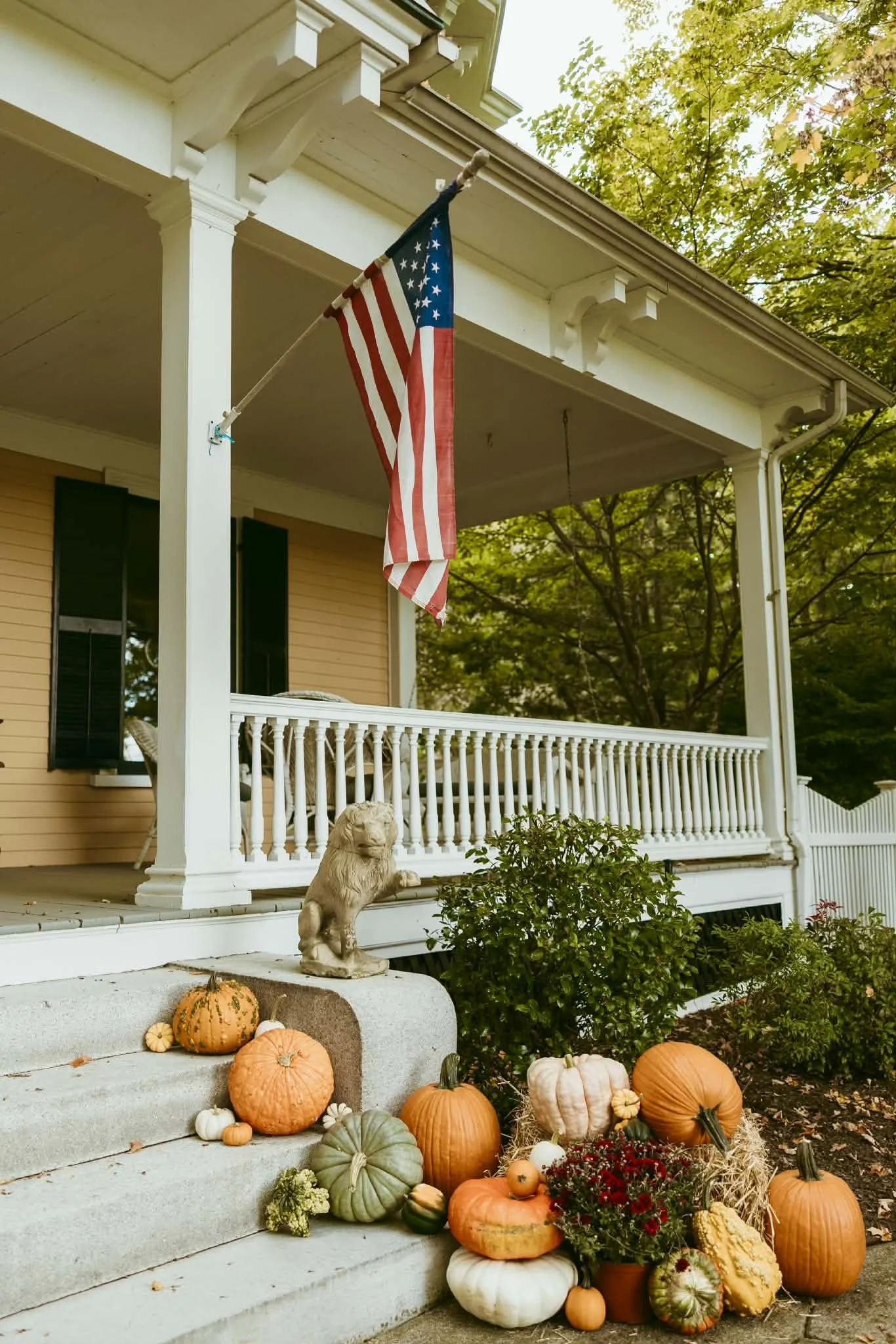 Front porch professionally decorated for fall with a U.S. flag, a lion statue, pumpkins, gourds, and fall flowers.