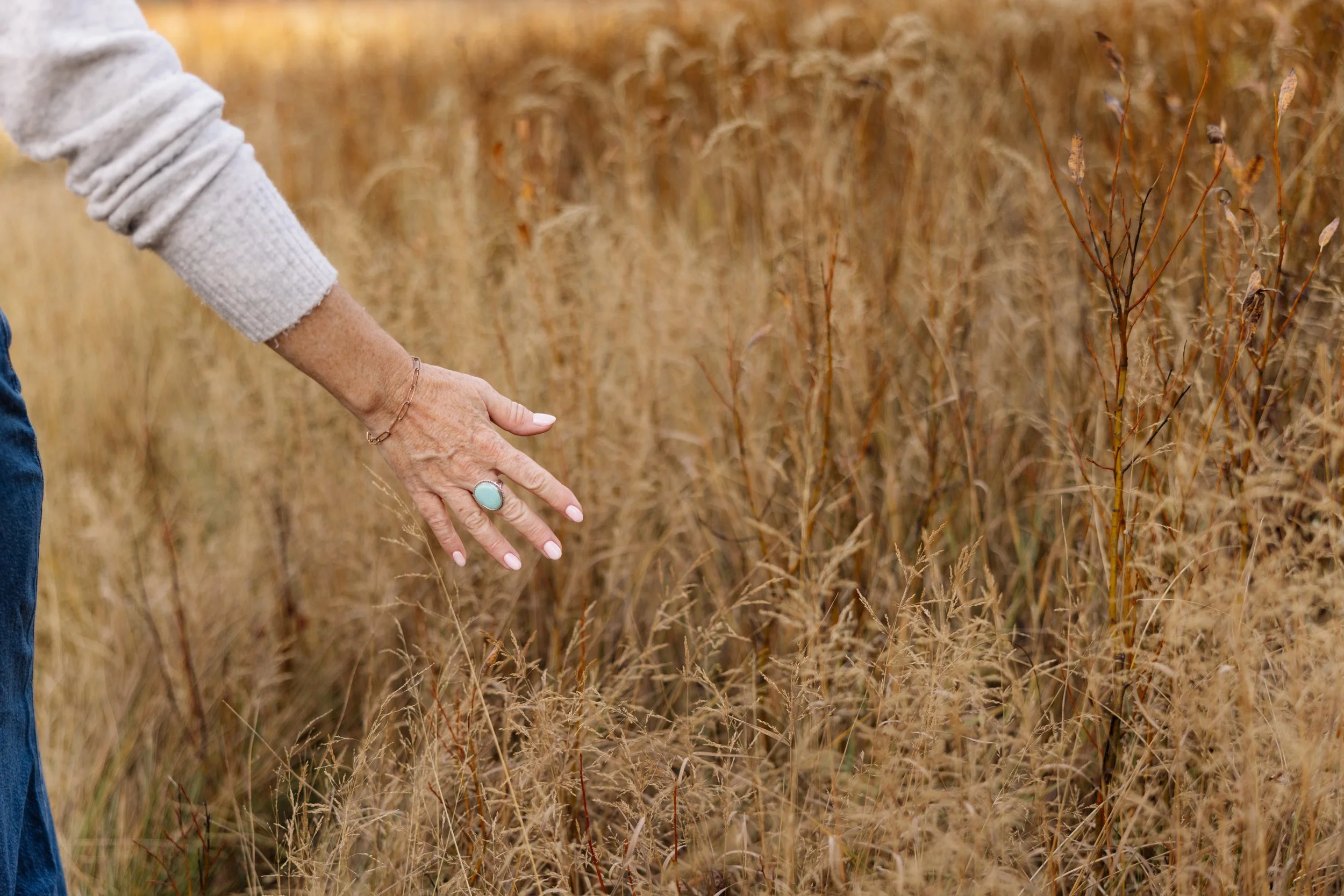 A woman extending her hand over a field of dry, tall grass, wearing a light-colored sweater and a turquoise ring.