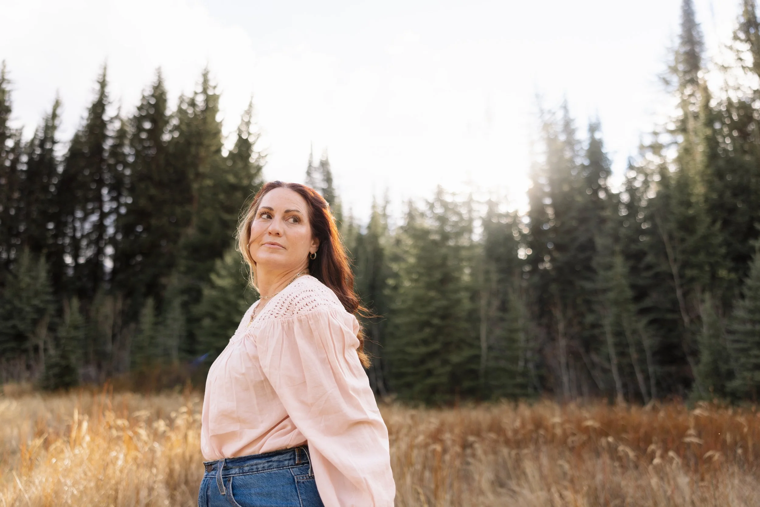 A woman with shoulder-length brown hair, wearing a light pink blouse and jeans, standing outdoors in a field with tall grass and trees in the background, lit by sunlight.
