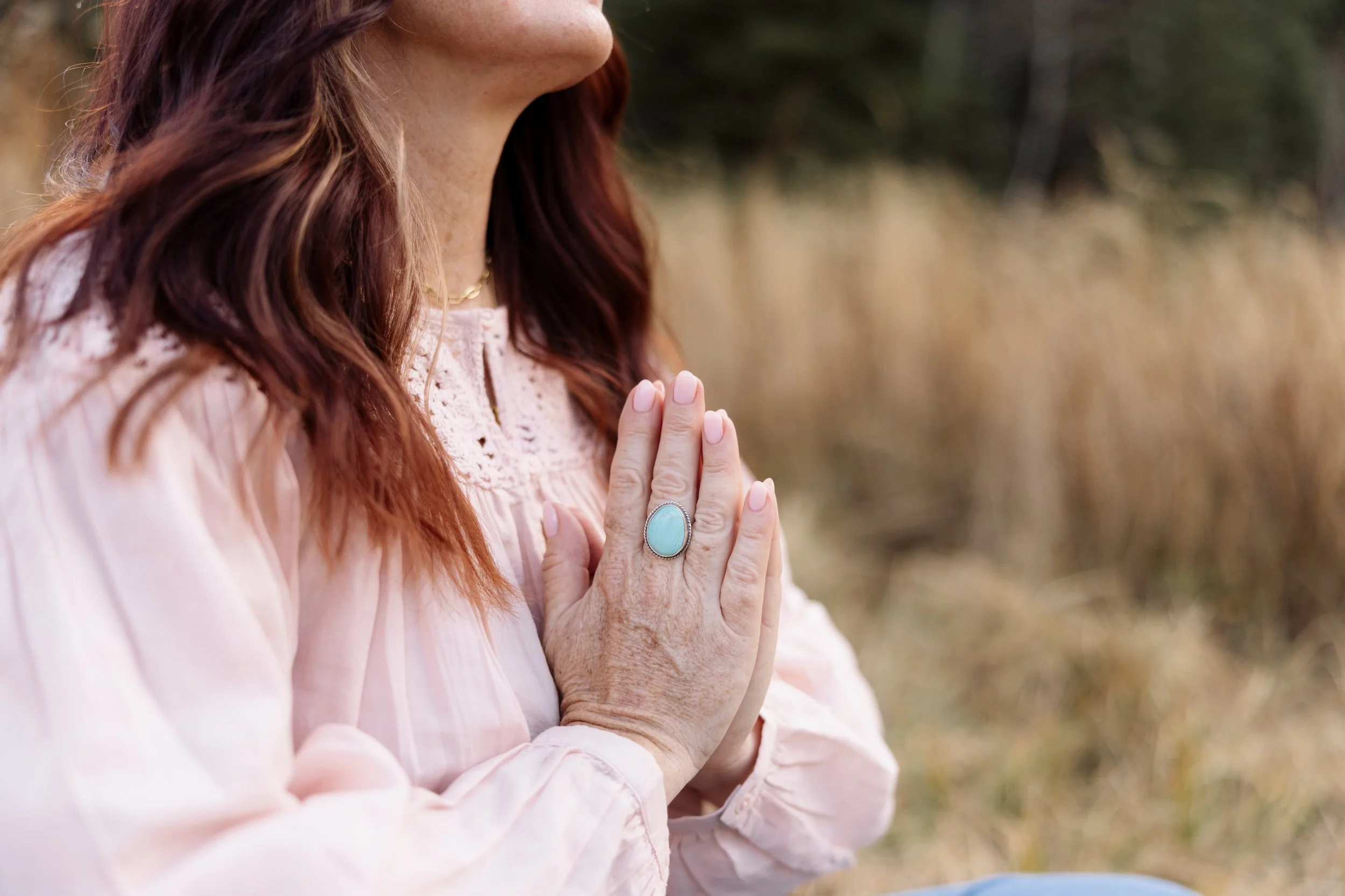 A woman with red hair is practicing meditation or prayer outdoors, with her hands pressed together in front of her chest. She is wearing a pale pink blouse and a large turquoise ring. The background shows blurred tall, dry grass.