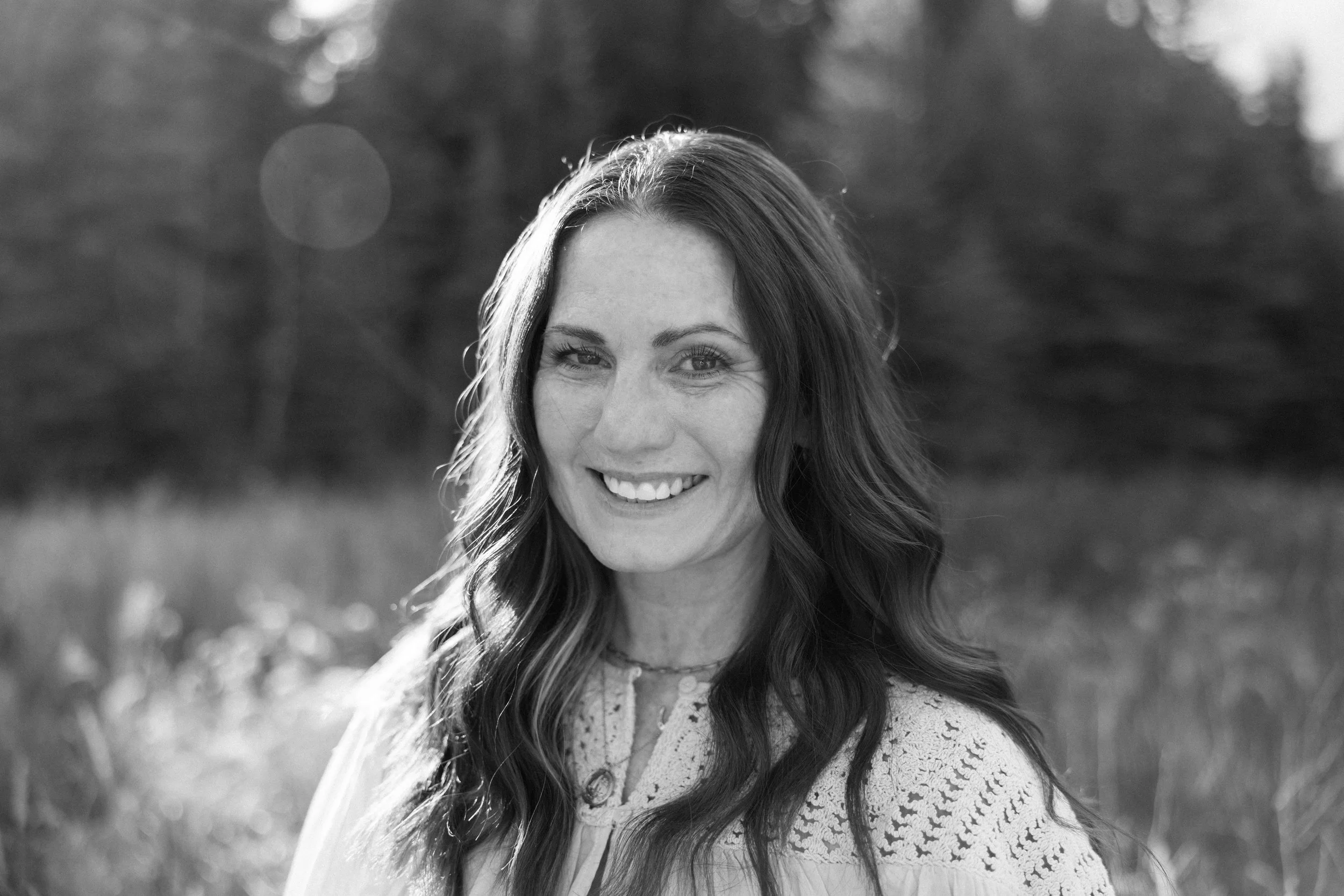 Black and white photo of a smiling woman with long wavy hair, wearing a light-colored top with lace details, standing outdoors with blurred trees and grass in the background.