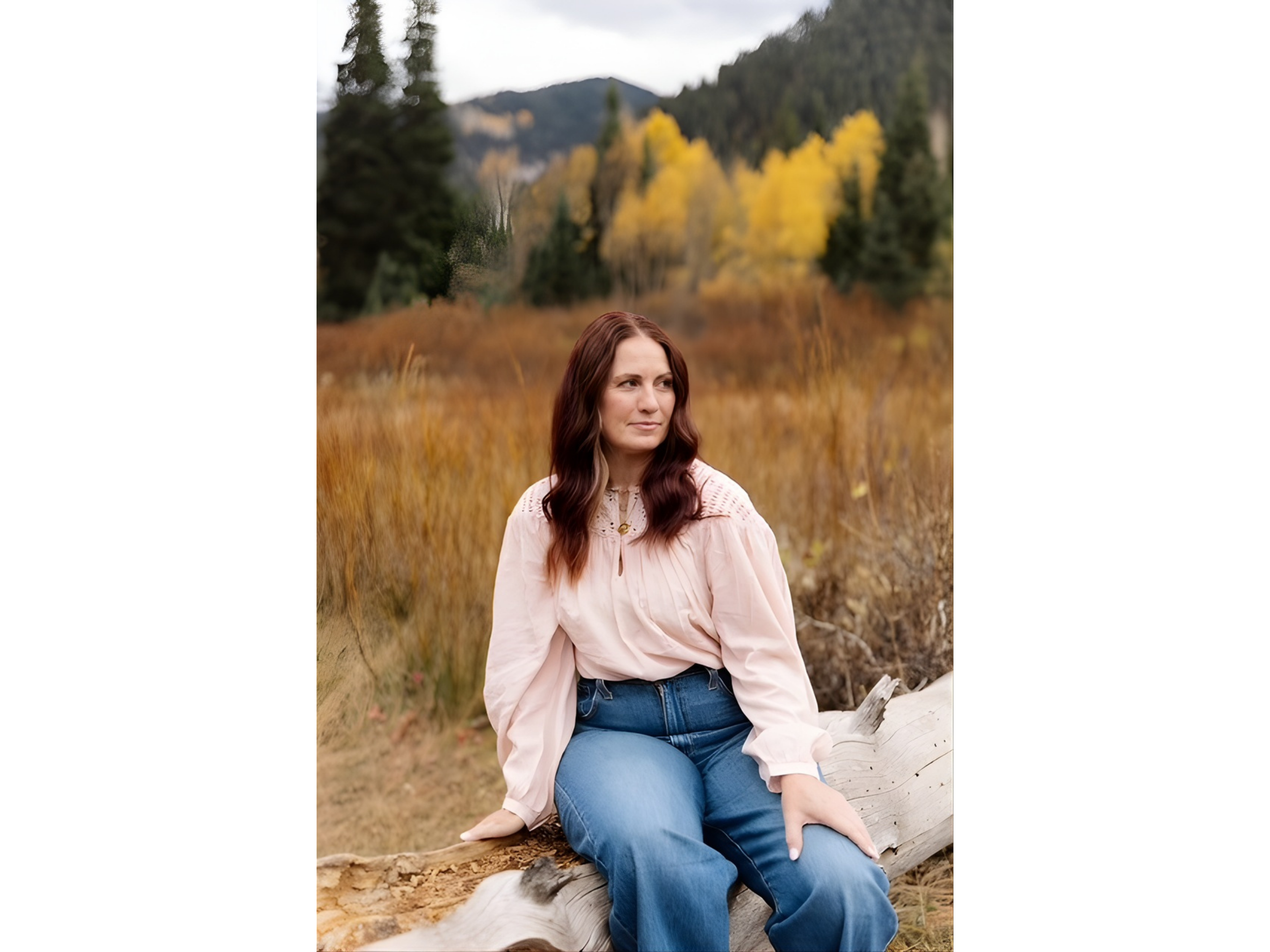 A woman with long brown hair sitting on a fallen log in a forested area with autumn foliage, wearing a light pink blouse and blue jeans.
