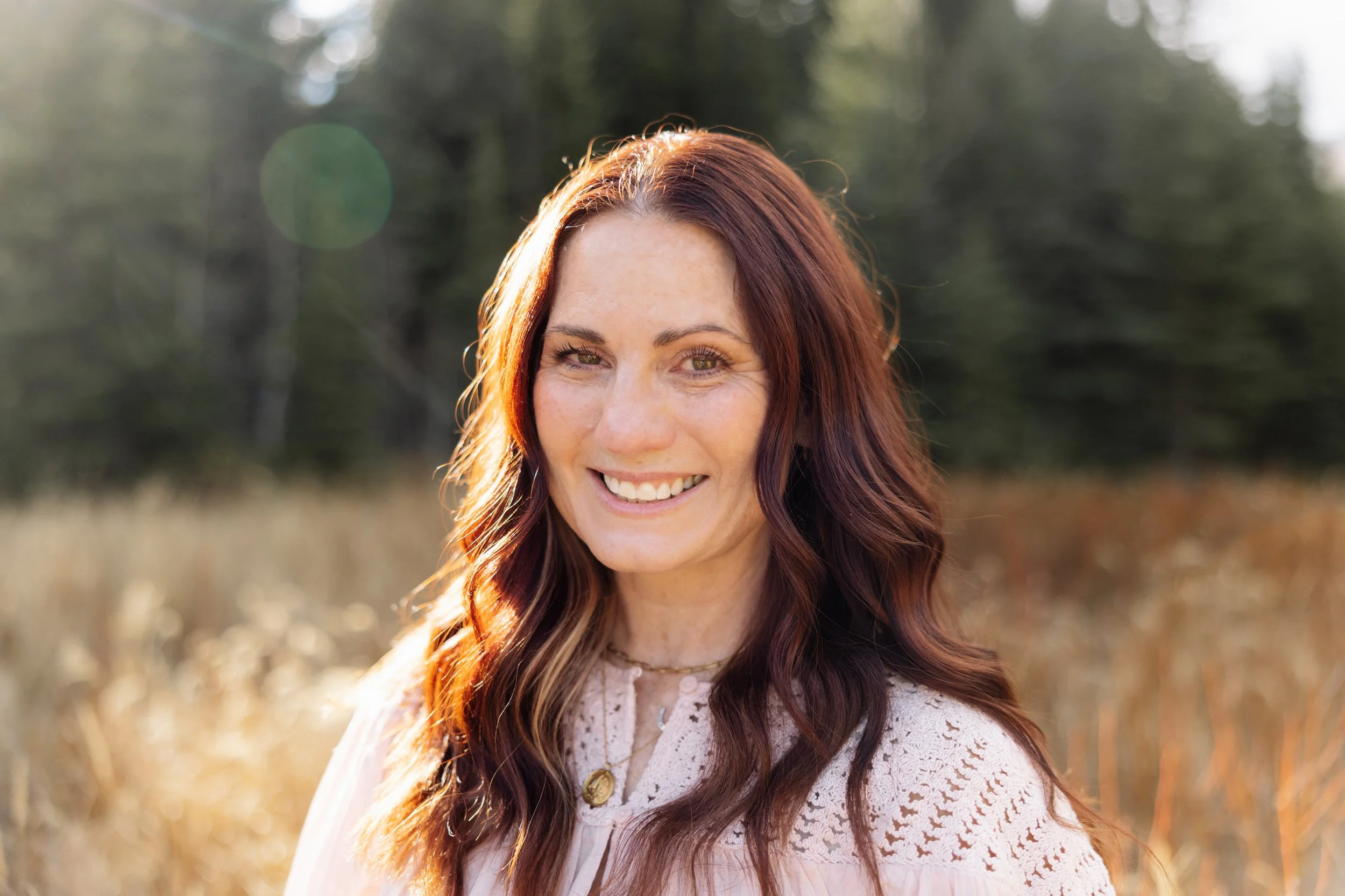 A woman with long wavy auburn hair smiling outdoors in a field with trees in the background during golden hour.