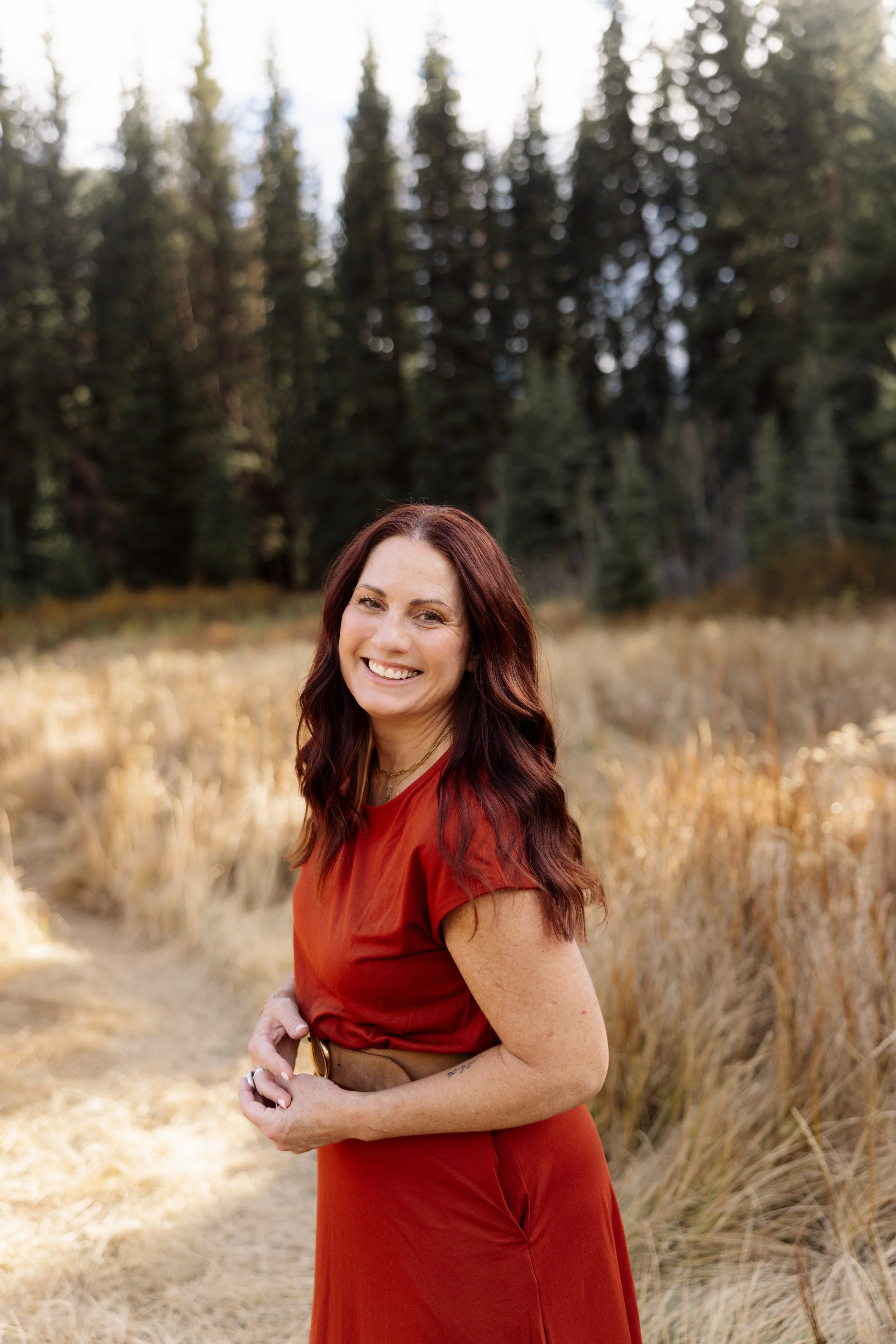 A woman with long red hair, wearing a red dress, smiling outdoors in a field with tall golden grass and a forest of pine trees in the background.