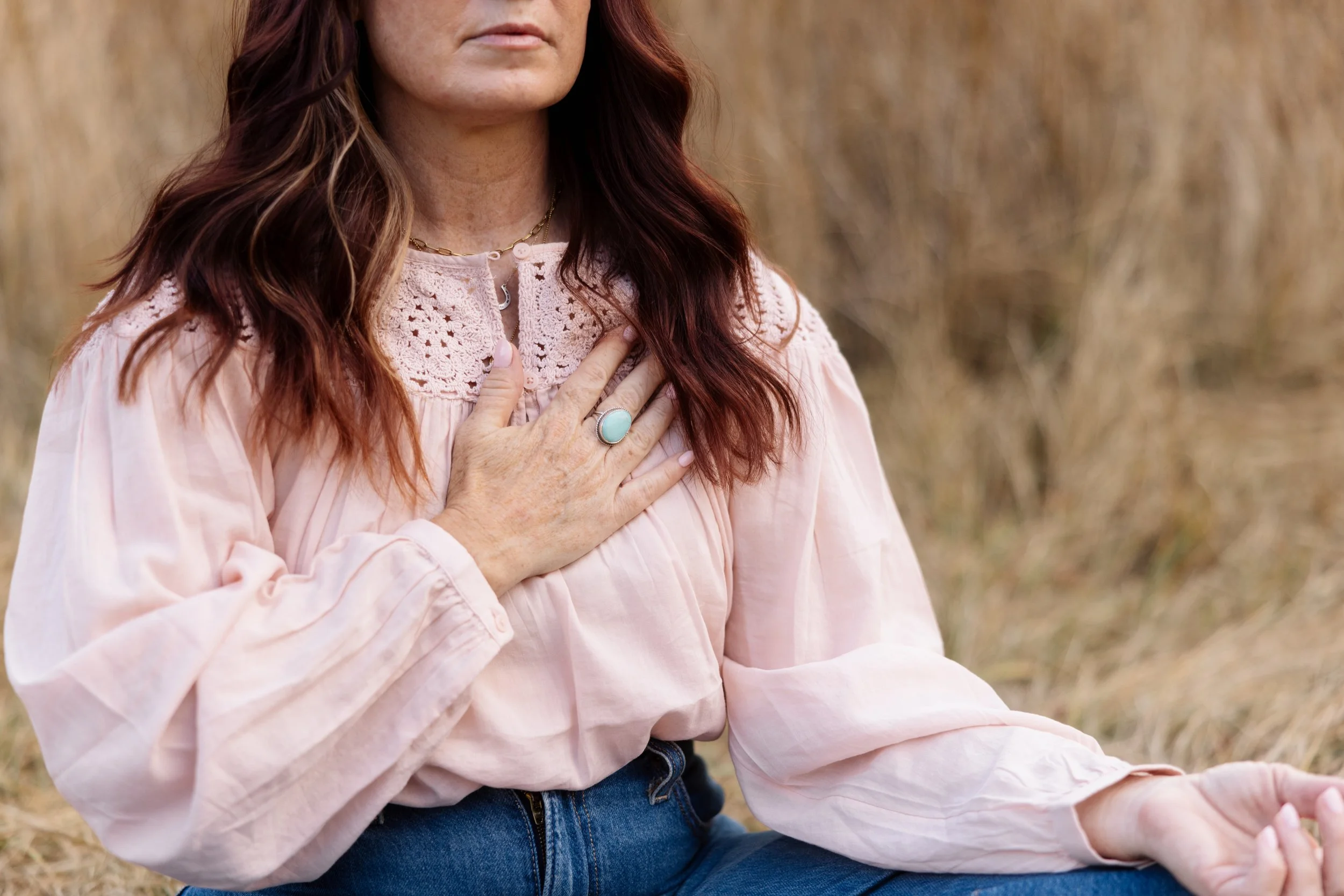 A woman in a pink blouse with crochet details, wearing a ring with a turquoise stone, sits outdoors on a grassy field, with her hand on her chest and her eyes closed.