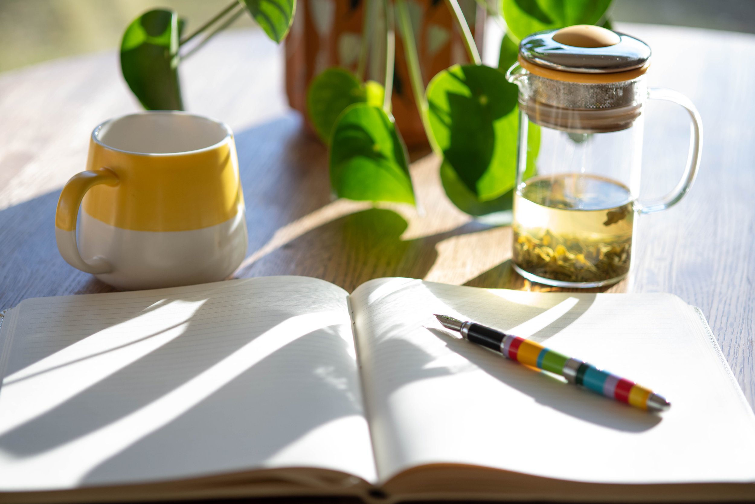 Open notebook with colorful pen, yellow mug, glass teapot with herbal tea, and green houseplants on wooden table, sunlight streaming in.