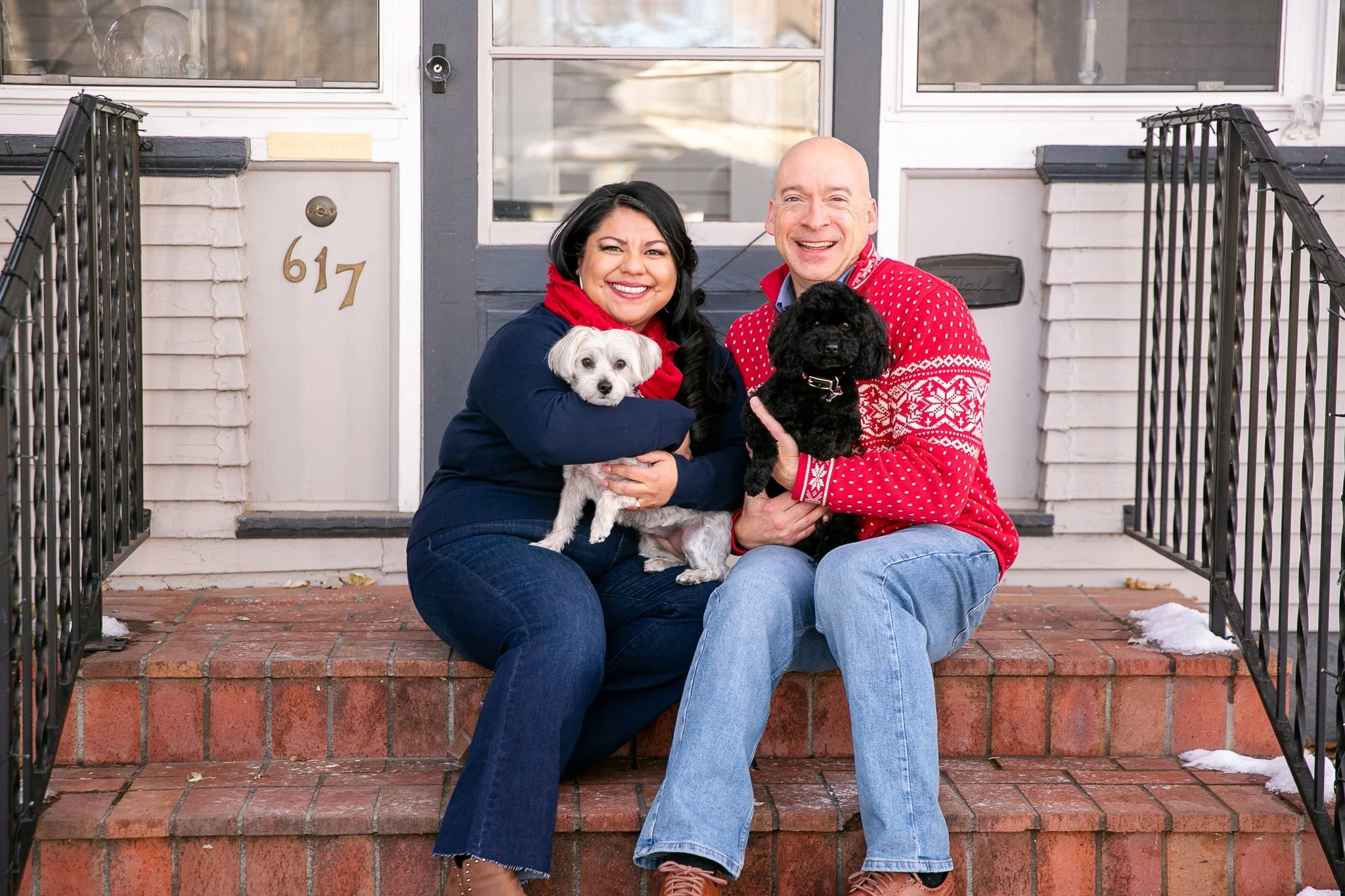 A smiling couple, a woman with black hair and a man with a bald head, sitting on their front porch with two small dogs, one white and one black, dressed in winter clothing, in front of a house with navy blue and white exterior walls.