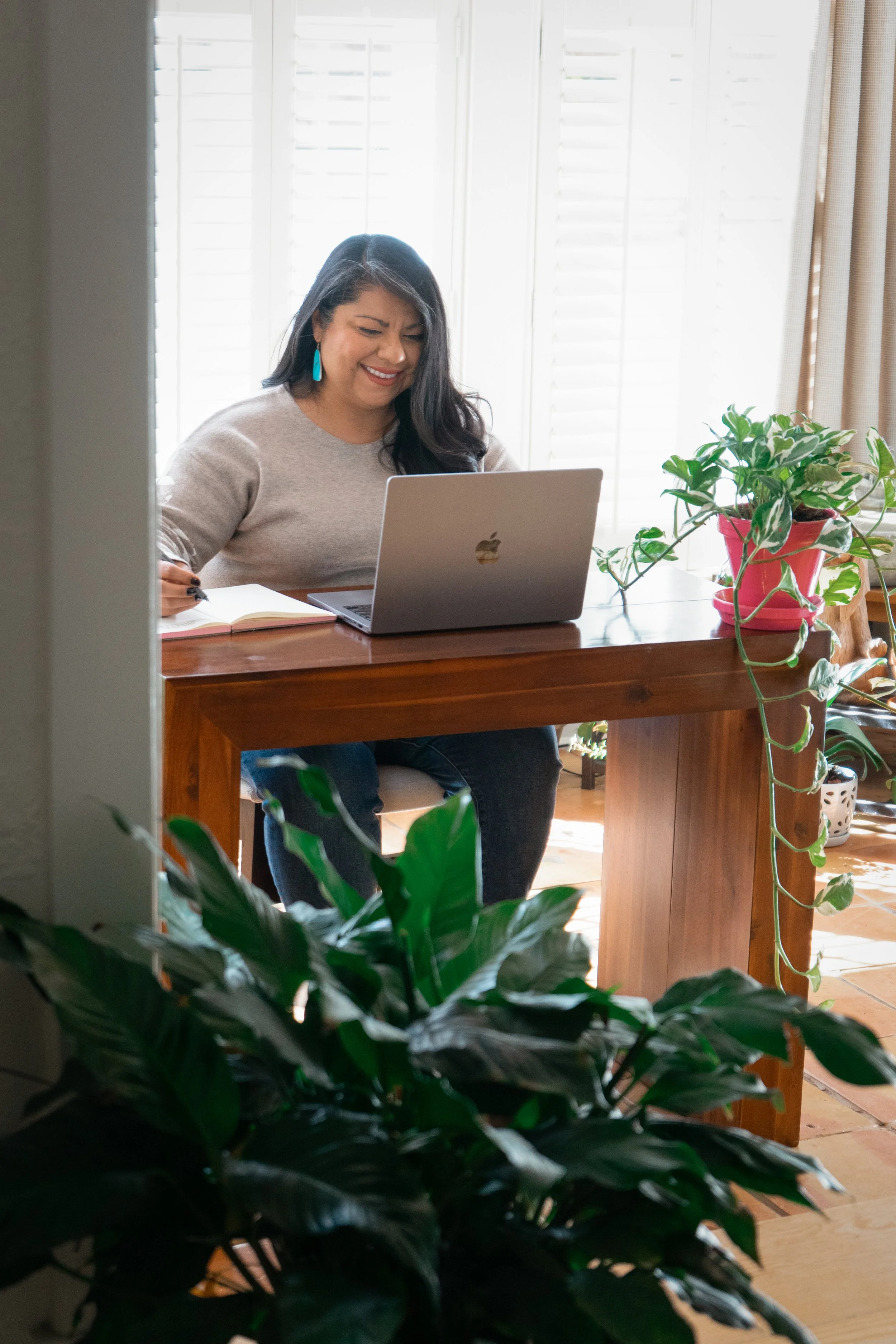 A woman with long dark hair, wearing turquoise earrings and a beige sweater, is sitting at a wooden table with a silver laptop, a notebook, and a pen, in a bright room with large white shutters on the windows and potted plants.
