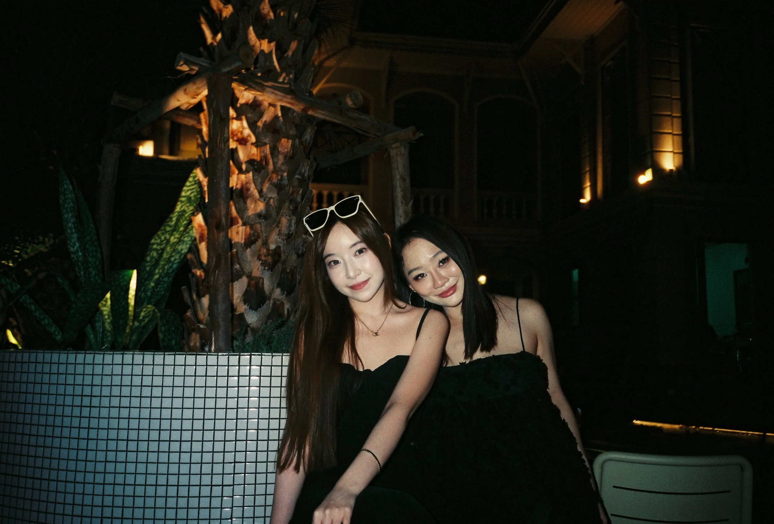 Two young women sitting close together and smiling at a nighttime outdoor event. They are dressed in black outfits and are sitting in front of a palm tree and a 185 year old colonial building.