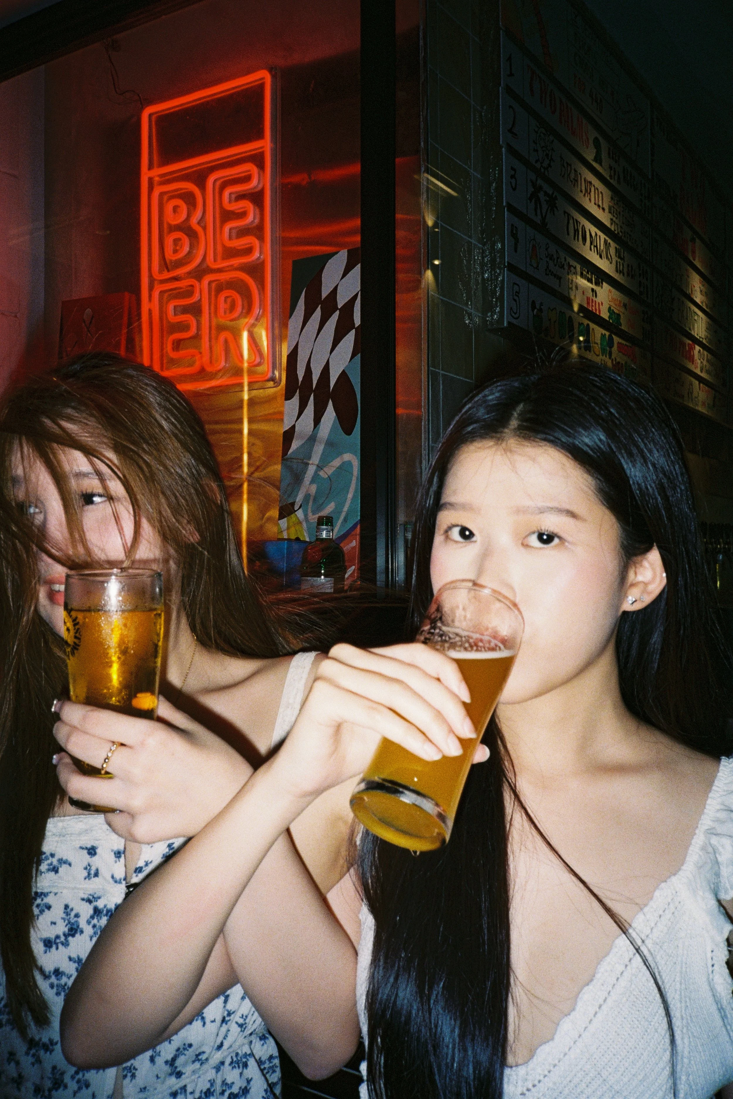 Two young women drinking beer at a bar or pub, with a red neon 'BEER' sign and colorful posters in the background.