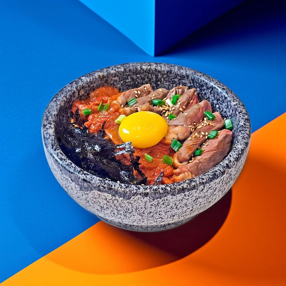 Bowl of bibimbap with raw egg yolk, sliced beef, vegetables, and sesame seeds on top, on a colorful background.